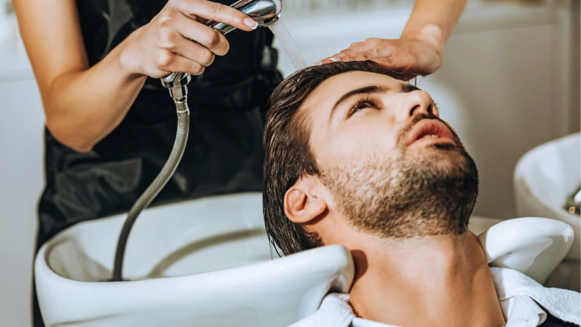 A man reclines at a salon sink, receiving a hair wash. A stylist gently rinses his hair, conveying relaxation and care in a bright, professional setting.