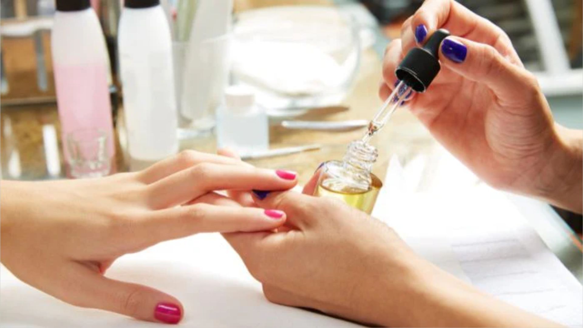Hands receiving a manicure, with one person applying cuticle oil using a dropper. The scene is relaxed, with nail care products in the background.