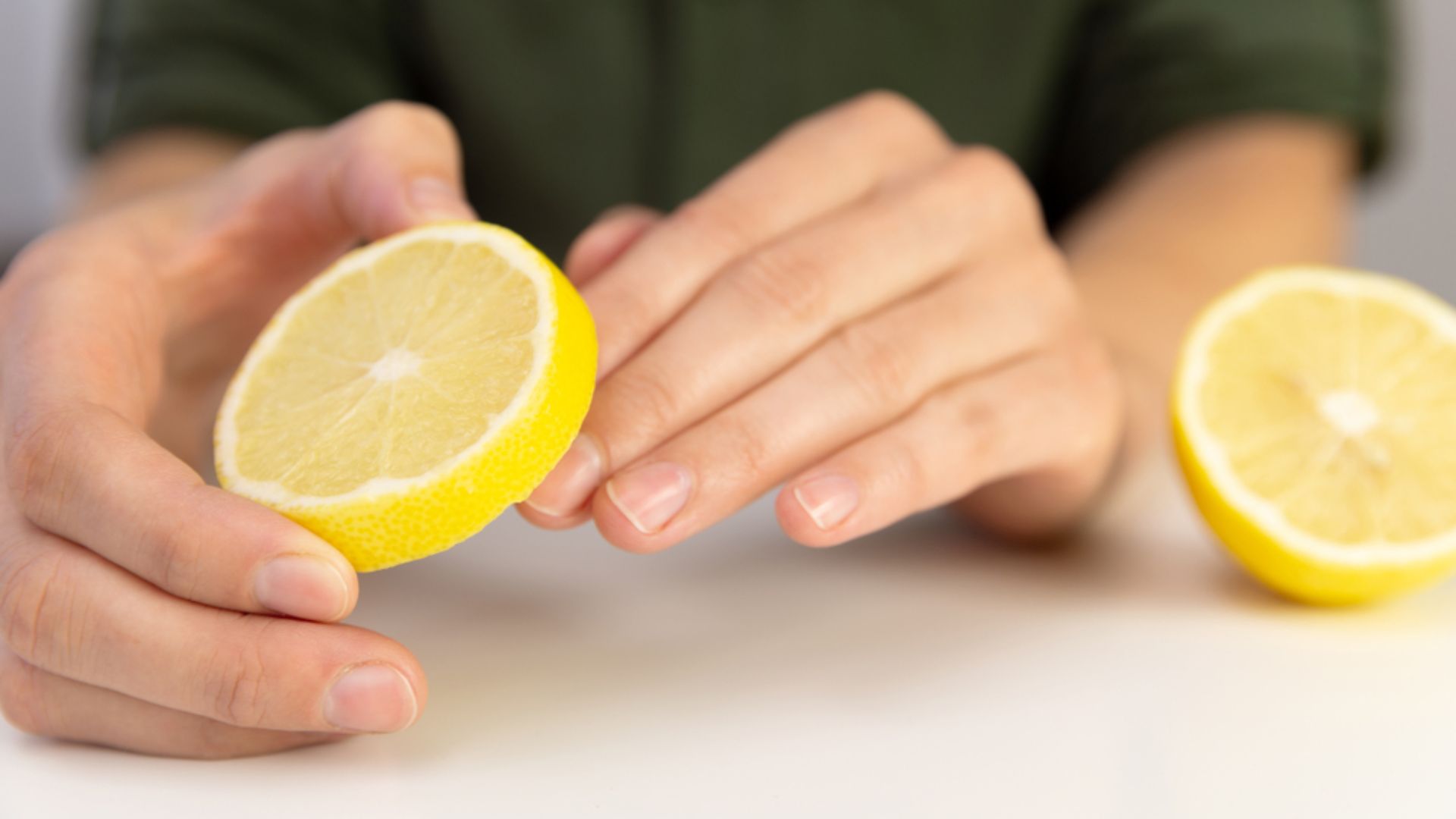 A person gently holds a fresh lemon half on a white surface, showcasing its bright yellow, juicy interior. Another lemon half lies nearby.
