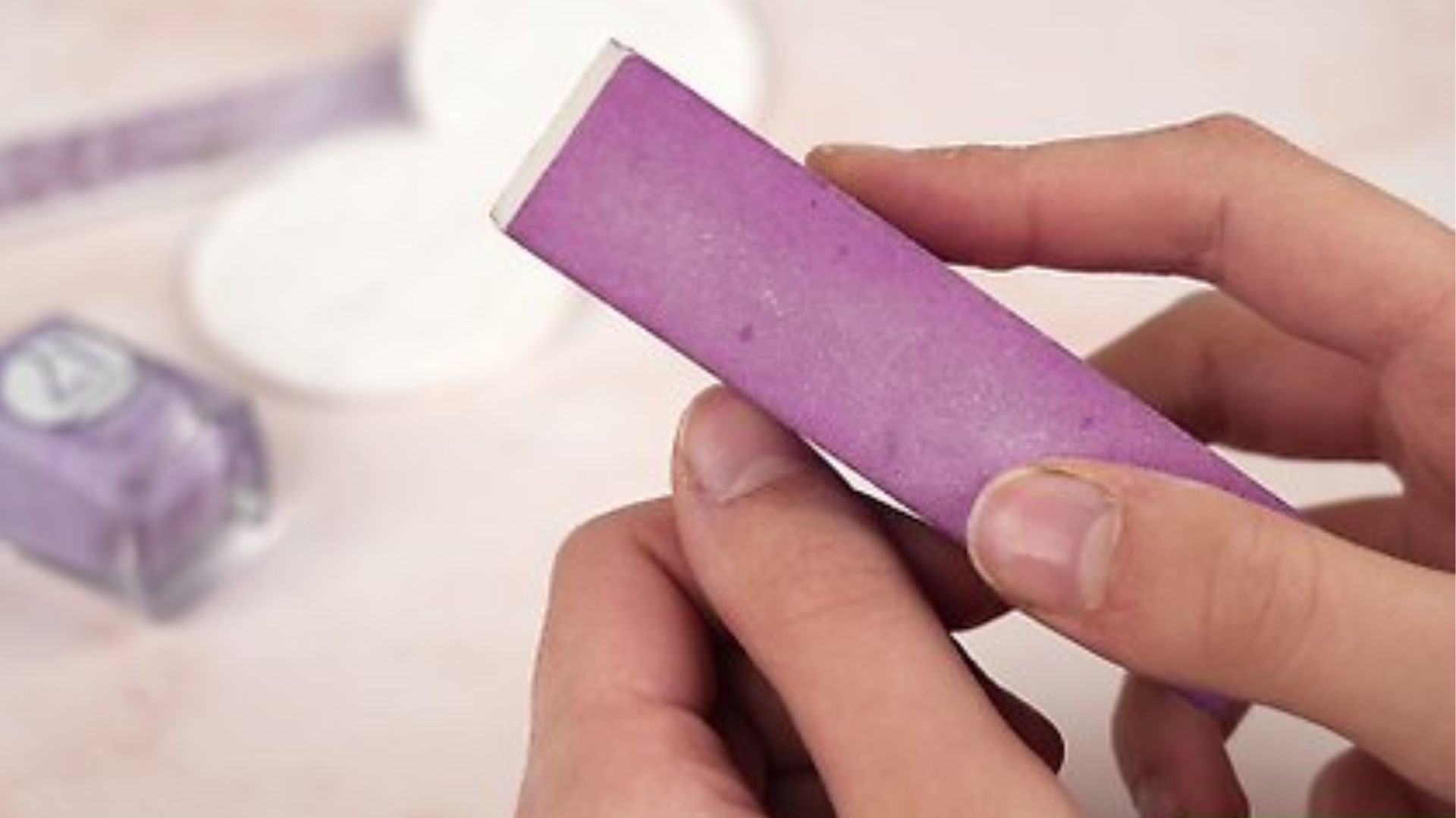 Hands holding a purple nail buffer, gently polishing a fingernail. In the background are blurred nail care items, conveying a calm self-care moment.