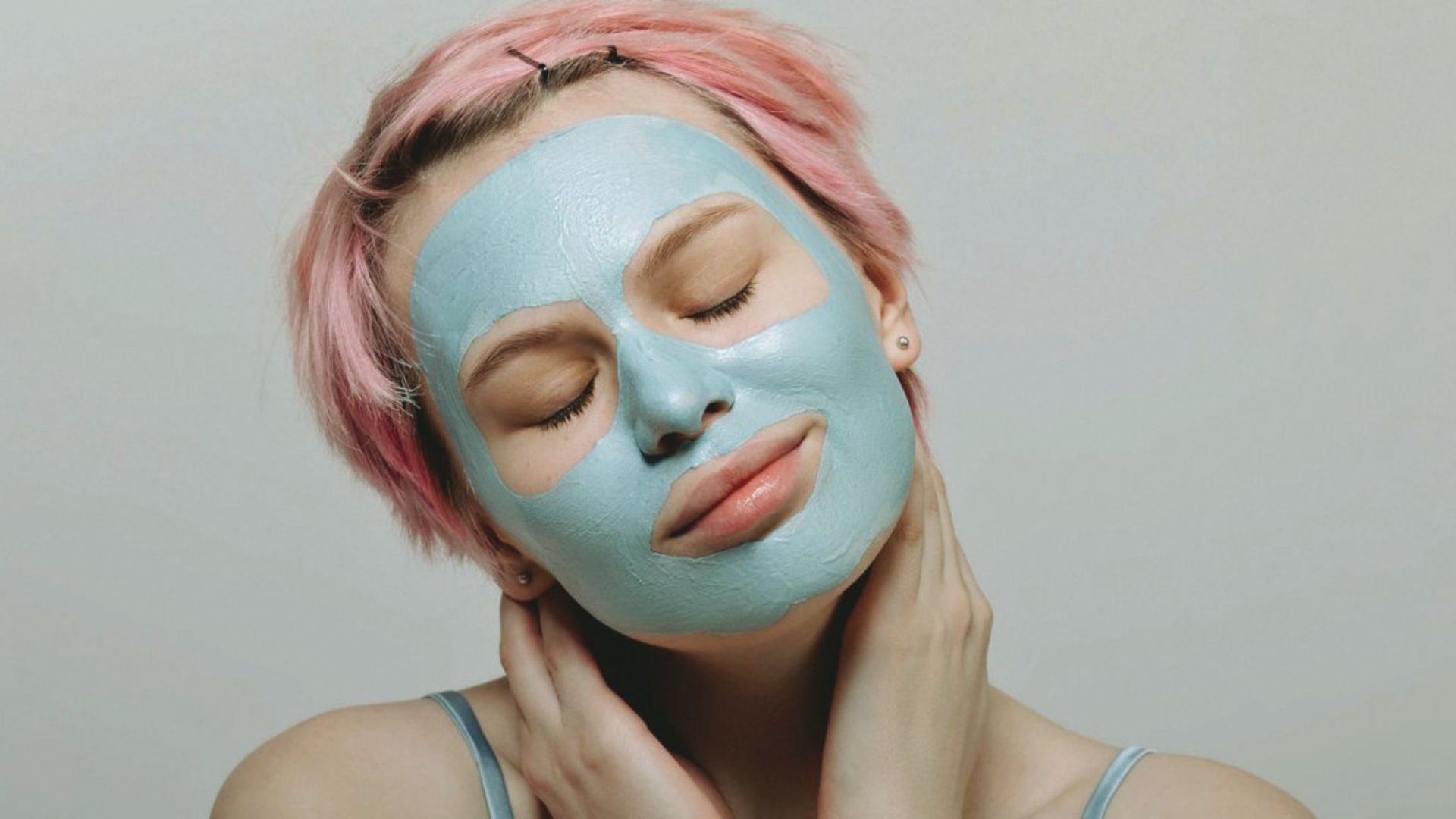 A woman with pink hair and a blue face mask gently holds her neck, eyes closed, expressing relaxation and serenity against a light gray background.