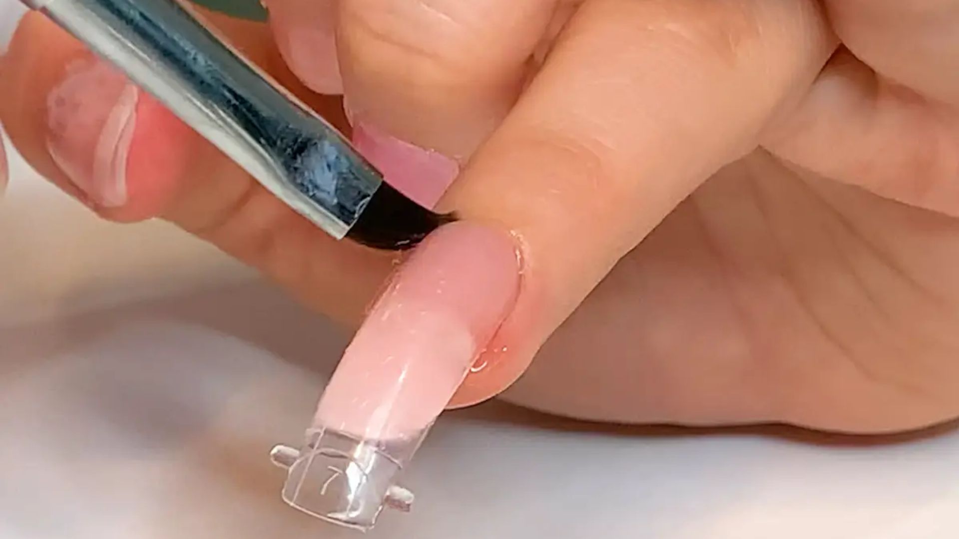 A close-up of a hand applying light pink acrylic to a long nail using a brush. A clear nail form is underneath, showing nail art in progress.