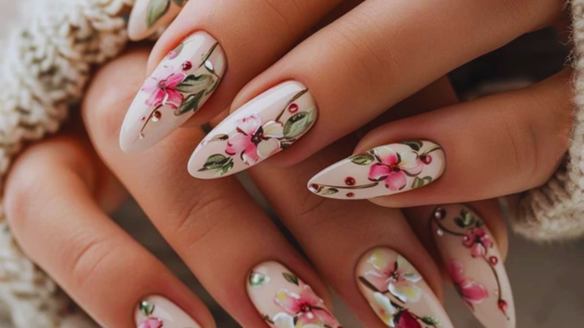Close-up of elegantly manicured hands with almond-shaped nails featuring intricate floral designs of pink flowers and green leaves, conveying a delicate and artistic tone.
