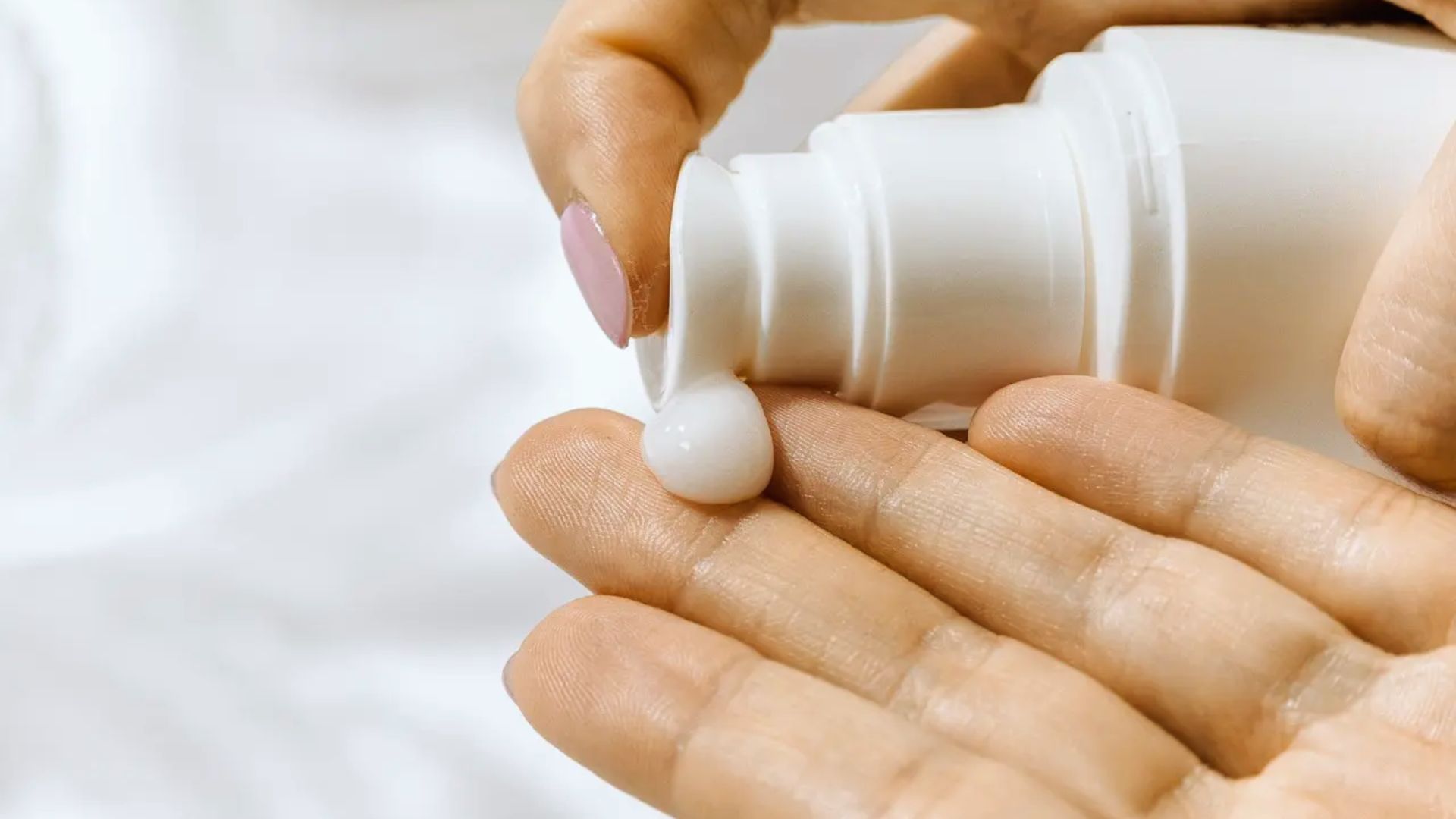 Close-up of a hand with light pink nails dispensing white lotion onto the palm from a pump bottle, set against a soft white background.