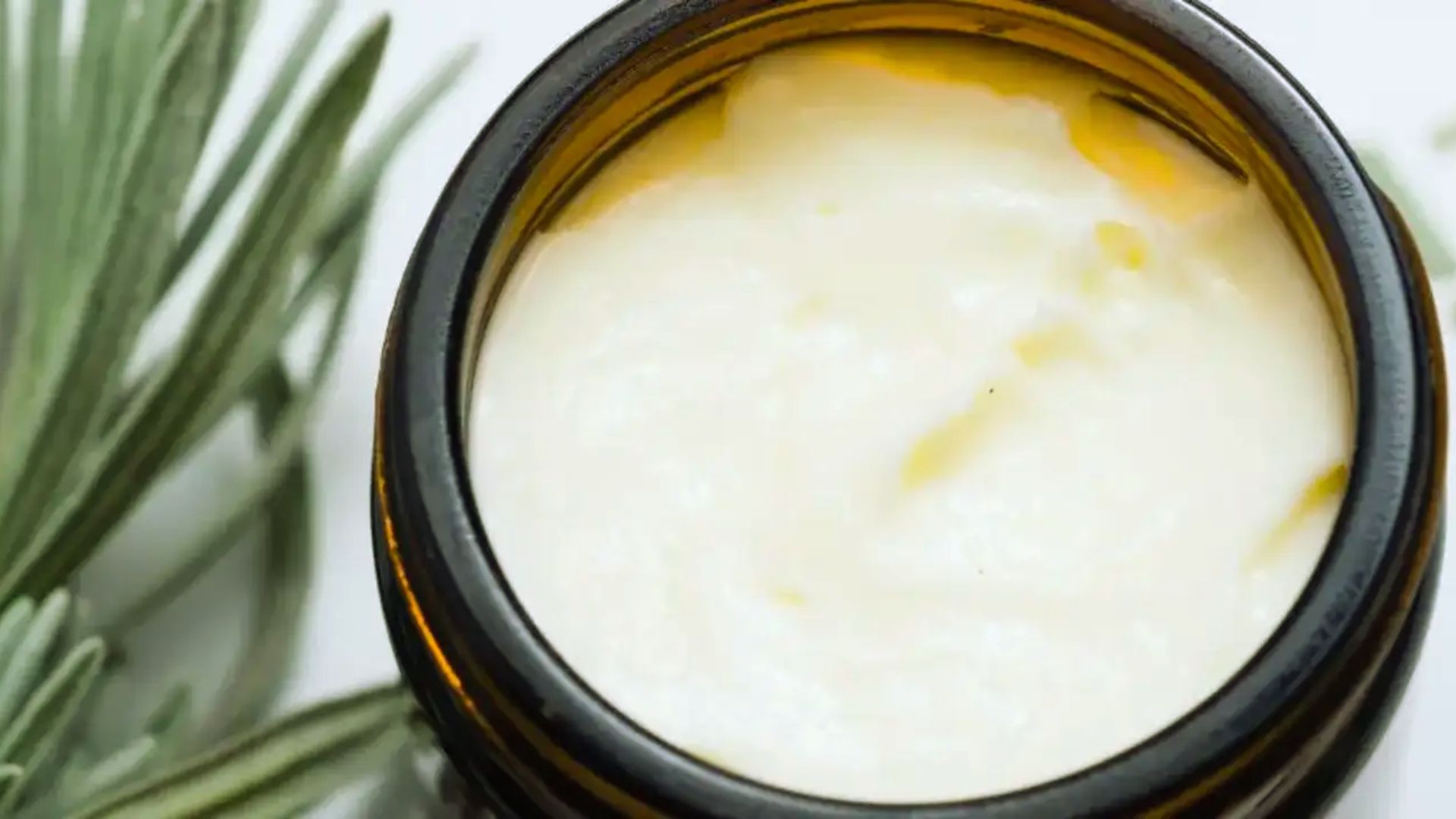 A jar of creamy, white ointment sits open, next to vibrant green leaves. The scene conveys a natural, soothing, and organic feel.