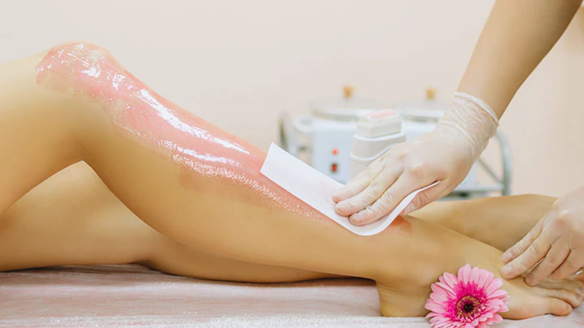 A person is getting their leg waxed in a spa setting. A gloved hand applies a wax strip, with a pink flower resting beside their feet. The atmosphere is calm and professional.