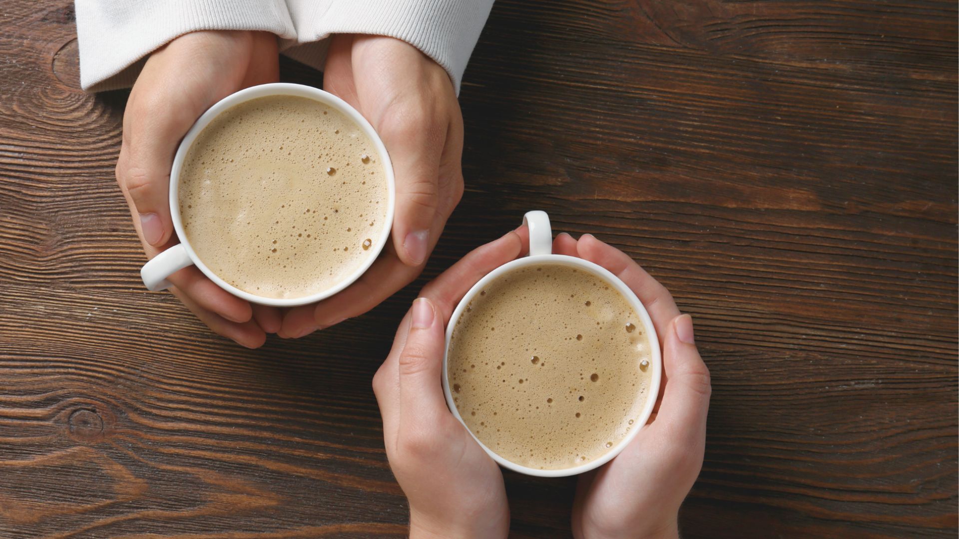 Two pairs of hands holding frothy coffee cups over a wooden table, suggesting warmth and companionship in a cozy setting.
