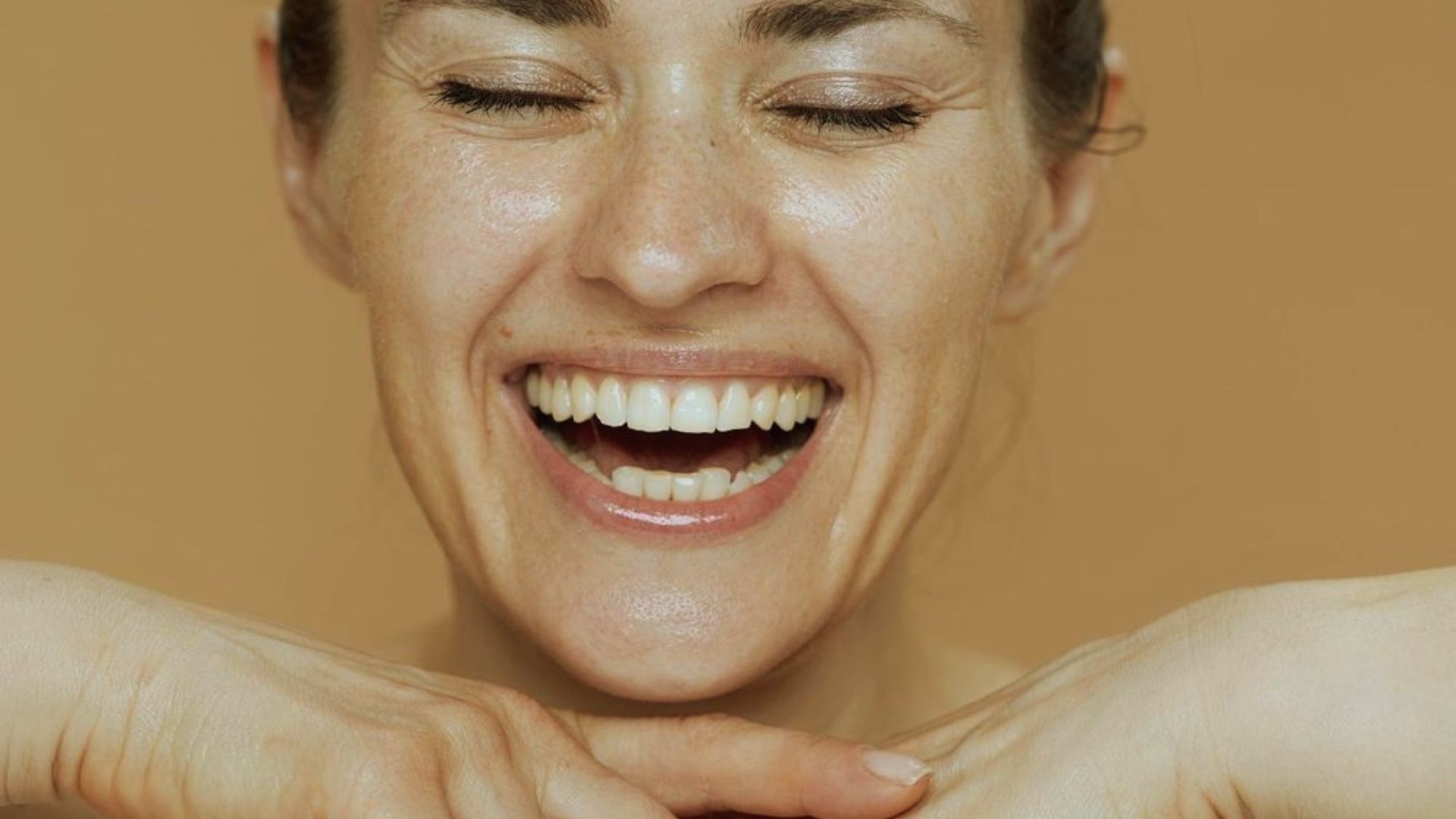 A woman with closed eyes and a joyous, wide smile rests her chin on her hands against a warm, brown background. Her expression conveys happiness.