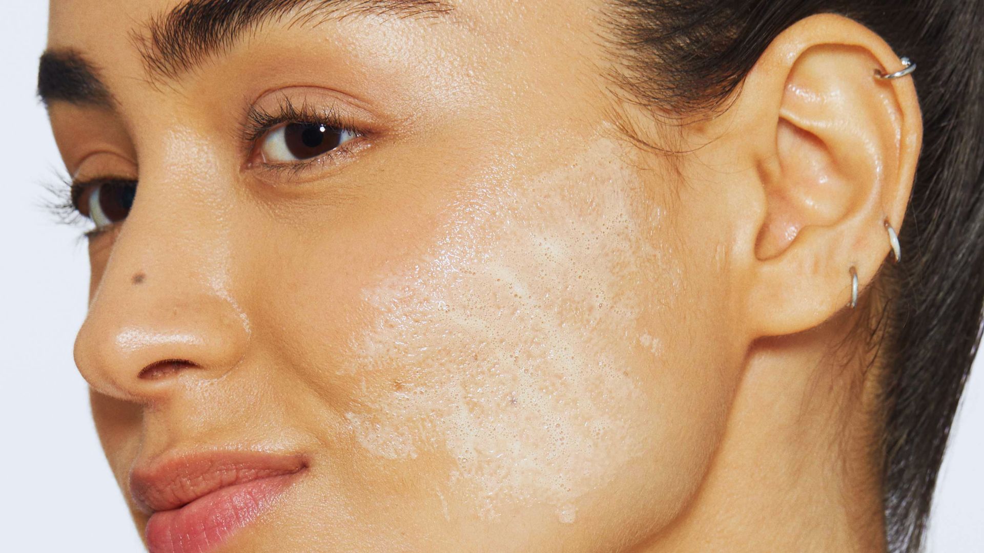 A close-up of a person's face with exfoliating cream applied on the cheek. Their expression is calm, showcasing smooth skin and multiple ear piercings.
