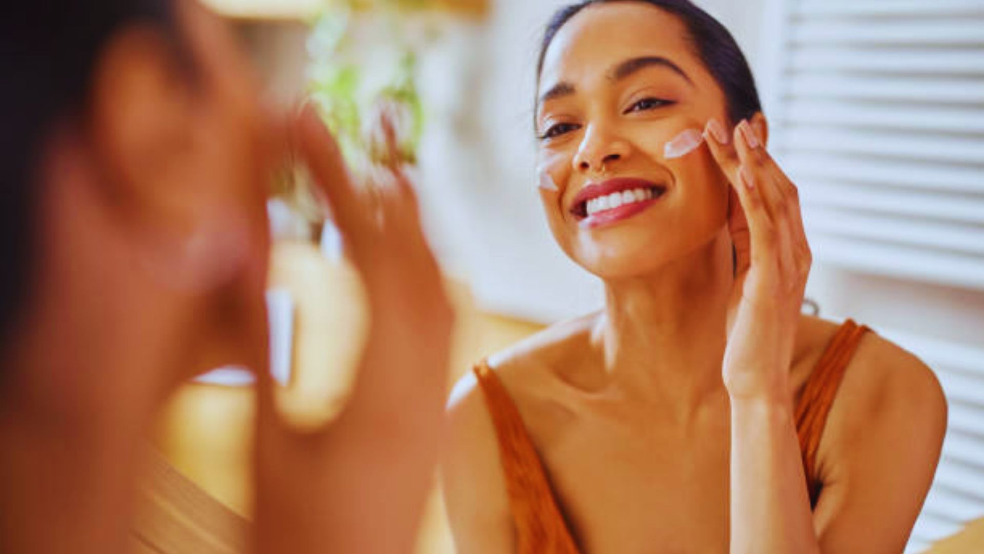A woman in a brown tank top smiles brightly while applying moisturizer on her cheeks, looking into a mirror. The scene is warm and inviting, conveying self-care.