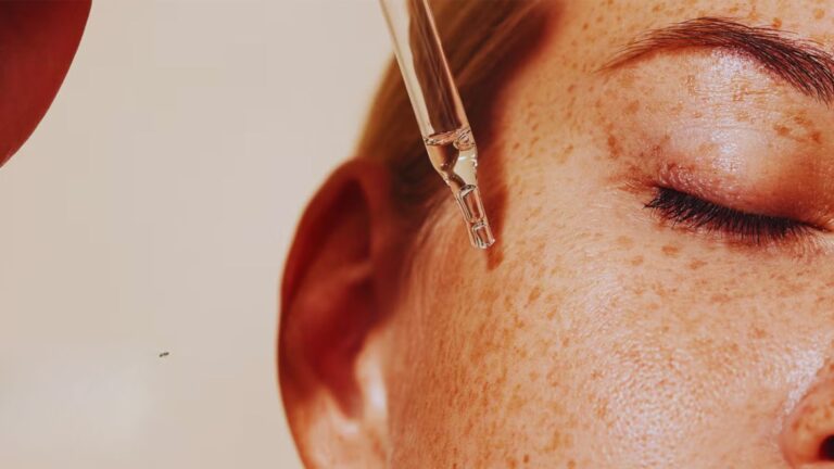 Close-up of a woman’s face with freckles, eyes closed, as a dropper applies serum to her skin. The image conveys a calm, soothing skincare routine.