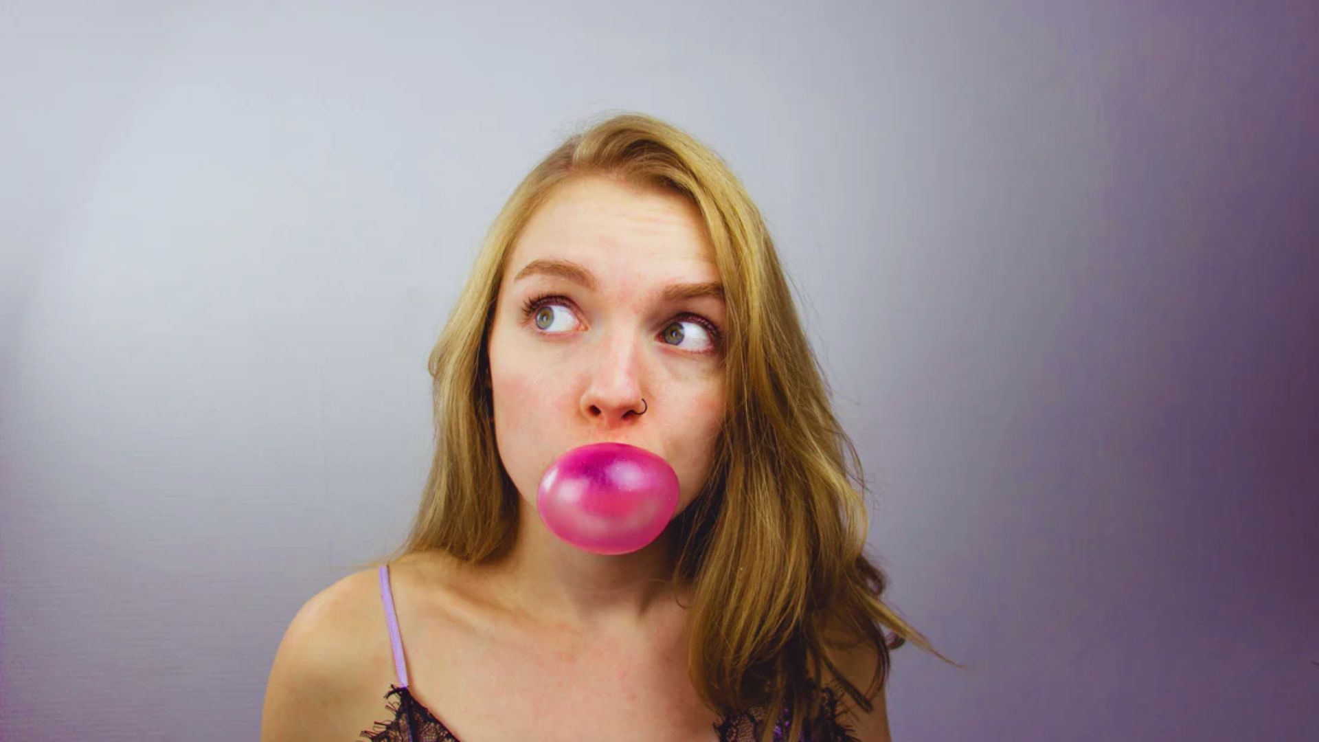 A woman with long blonde hair blows a pink bubblegum bubble, looking playfully to the side. She wears a casual top against a plain background.