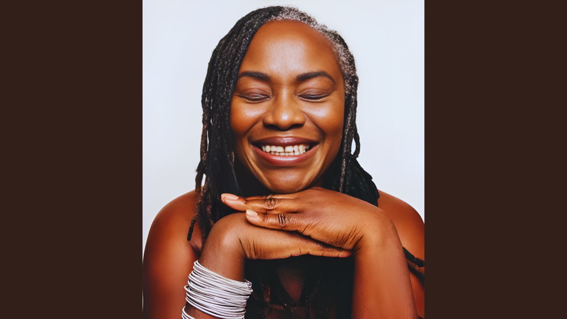 Smiling person with braided hair and closed eyes, resting their chin on hands. They wear silver bangles and convey warmth and joy against a plain background.