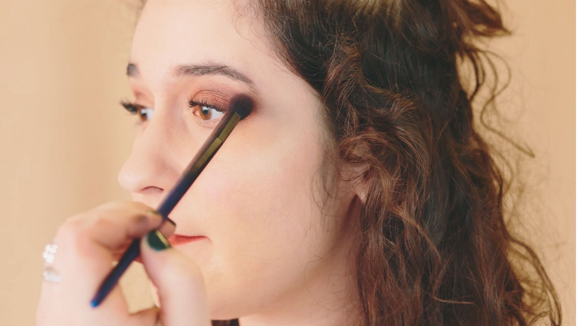 A person with curly hair receives a professional makeup application, focusing on blending eyeshadow. The setting conveys a calm and polished atmosphere.