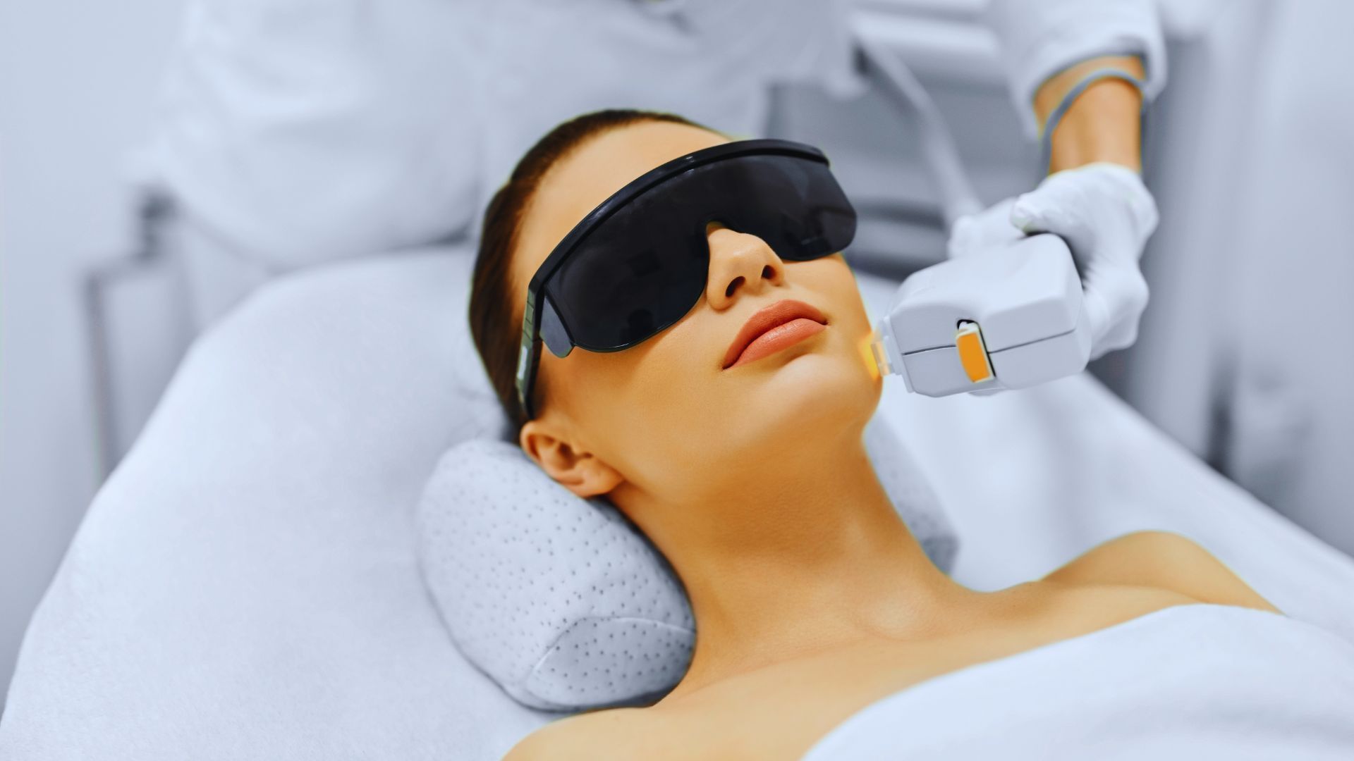 Woman receiving a facial laser treatment wearing protective goggles. A gloved hand holds the laser device, creating a clinical, calm atmosphere.