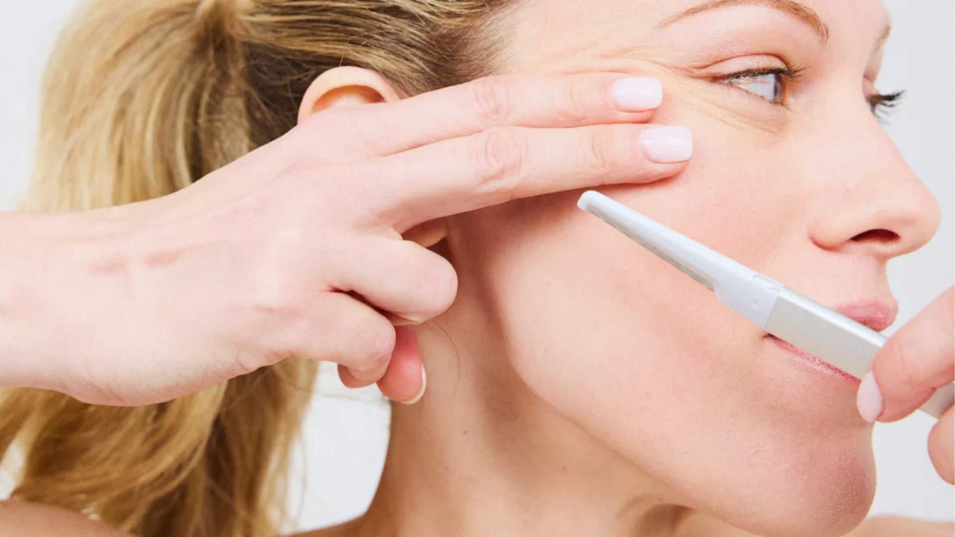 A woman gently holds her cheek taut with two fingers while using a white facial razor. The background is plain, emphasizing a skincare routine.