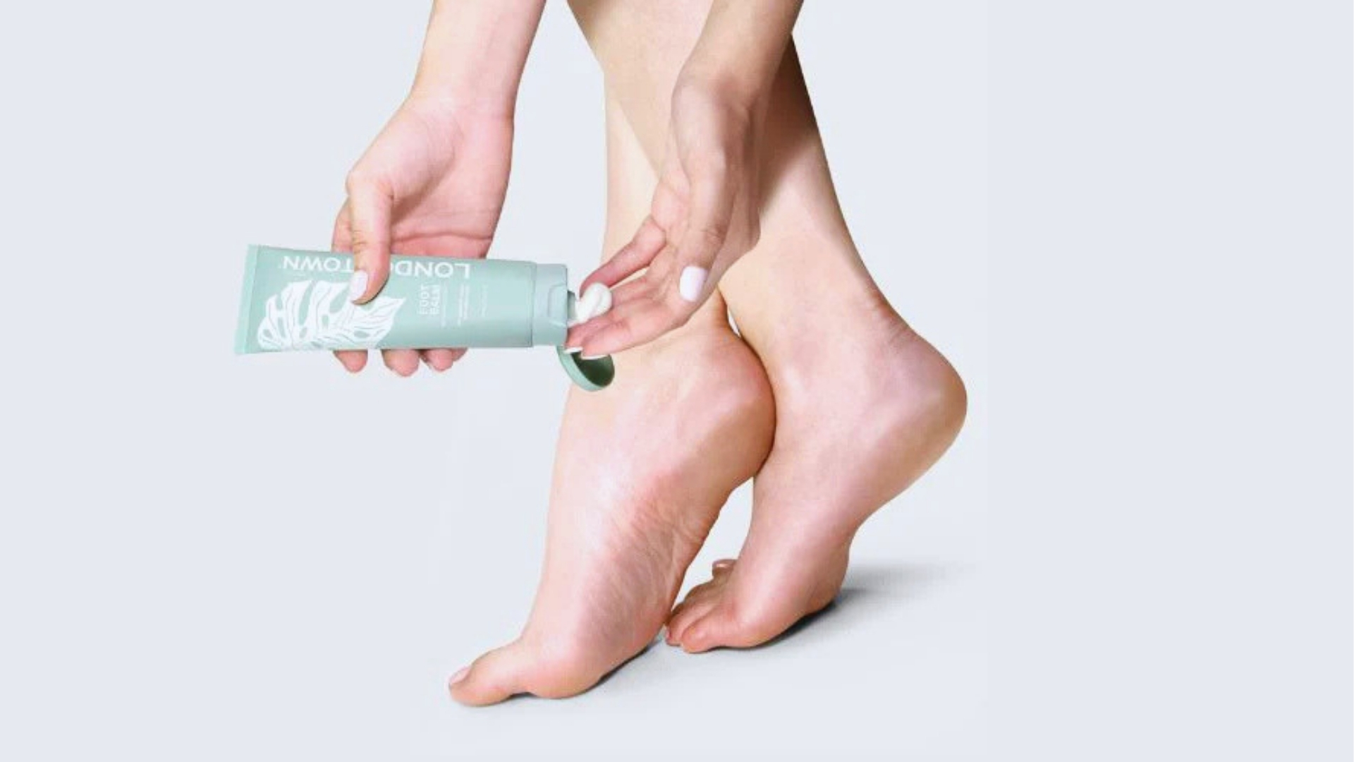 Hands applying lotion from a mint green tube onto bare, smooth feet against a light background, conveying a sense of self-care and relaxation.