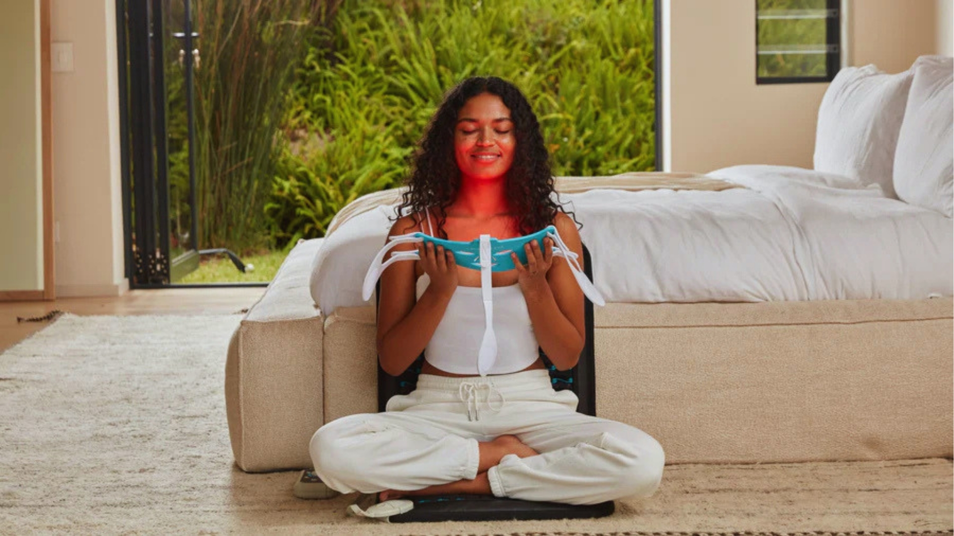 A woman sits cross-legged and relaxed in a bright room, holding an eye mask. Sunlight streams in through a window, and a neat bed is visible behind her.