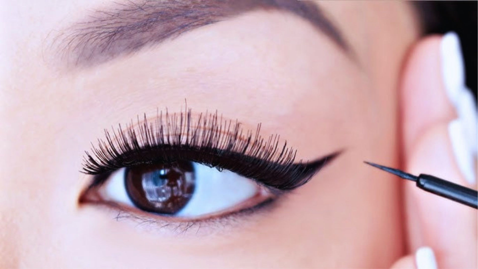 A close-up of an eye with bold, expertly applied black eyeliner and false lashes. The eyebrow is neatly shaped, and a hand holds a fine-tip eyeliner.