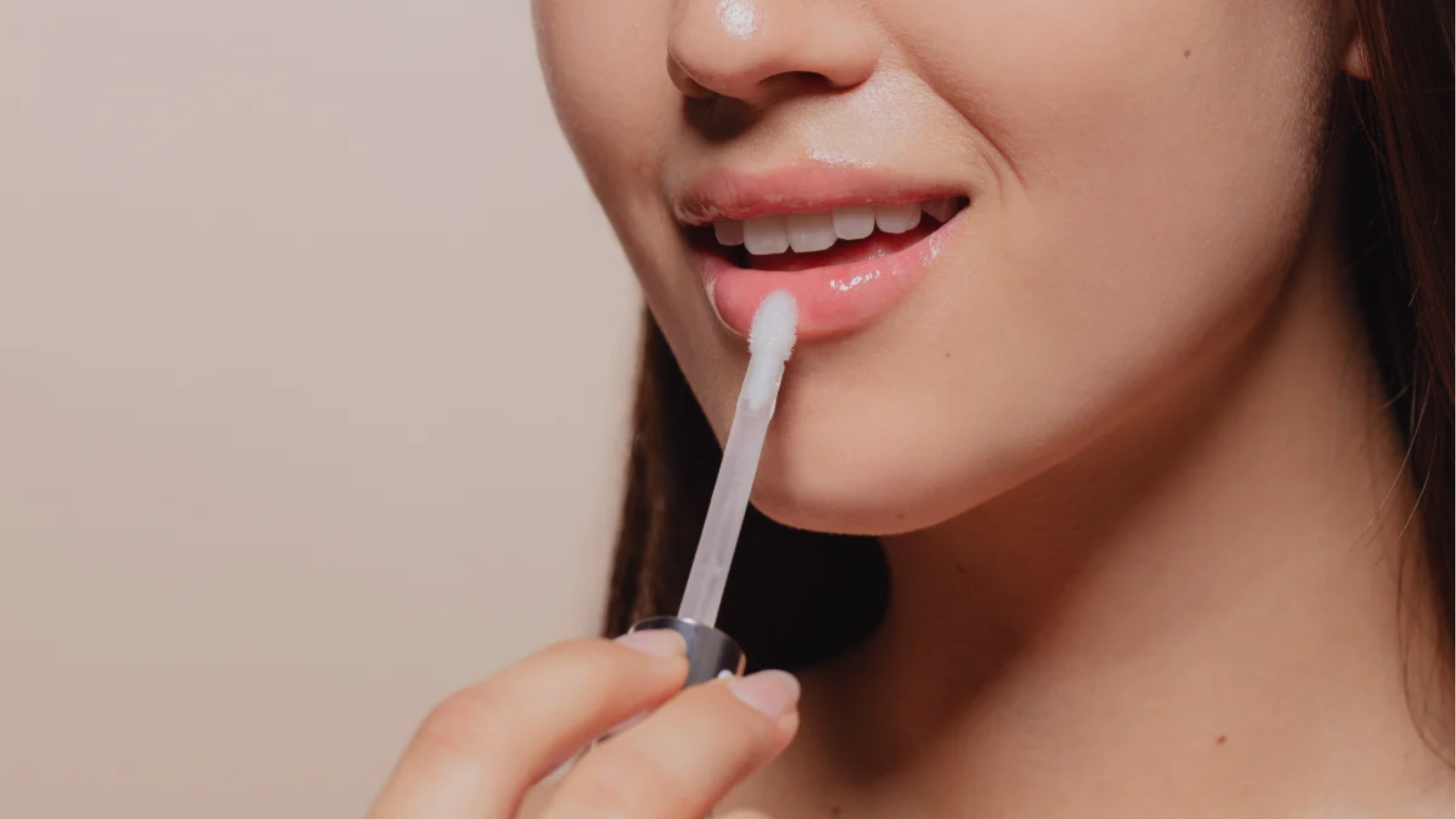 Close-up of a person applying clear lip gloss with an applicator to their lips. The background is neutral, highlighting smooth skin and a gentle expression.