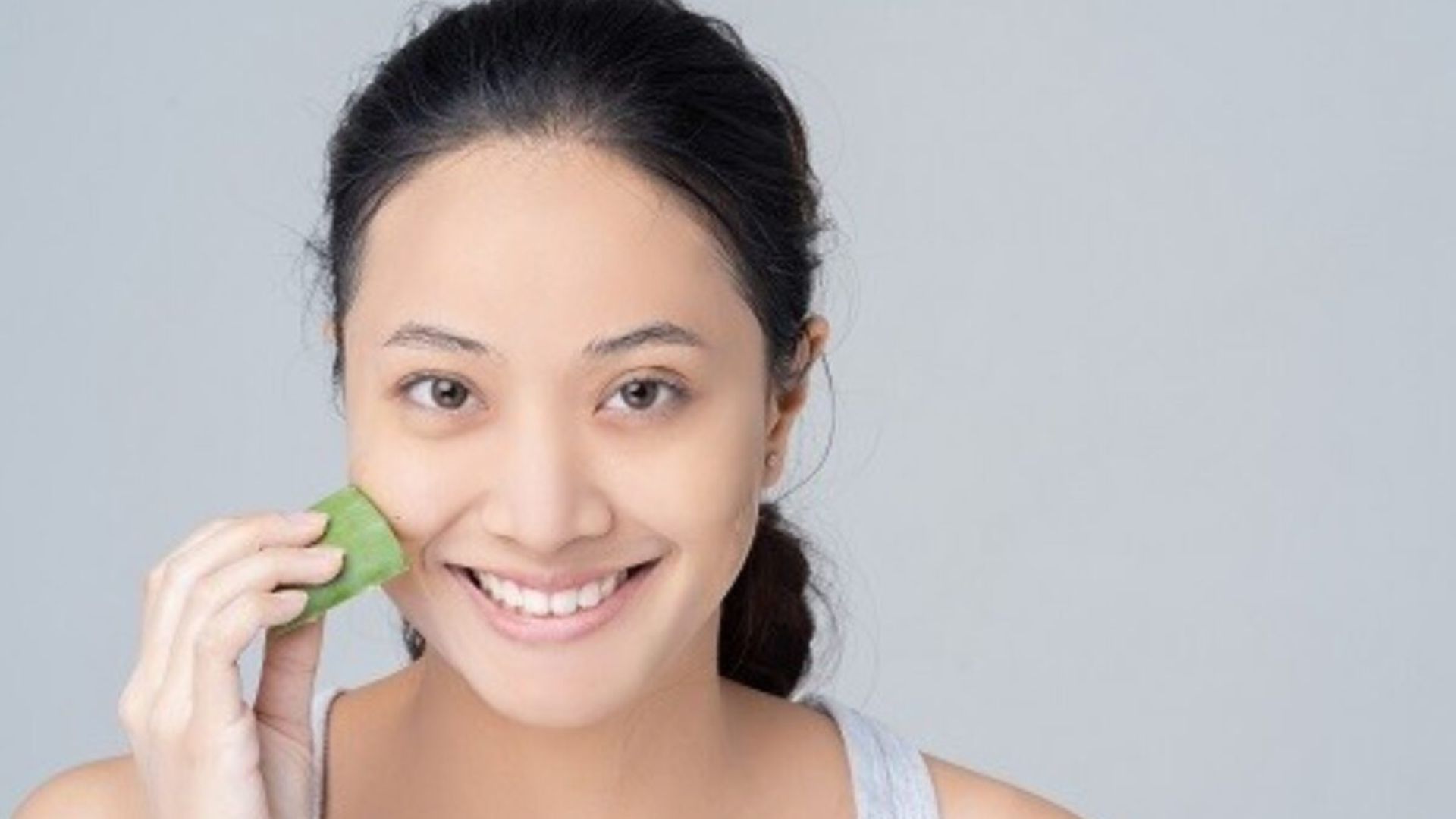 Smiling person gently rubs aloe vera on their cheek, conveying freshness and skincare. The background is light gray, creating a calm and soothing vibe.
