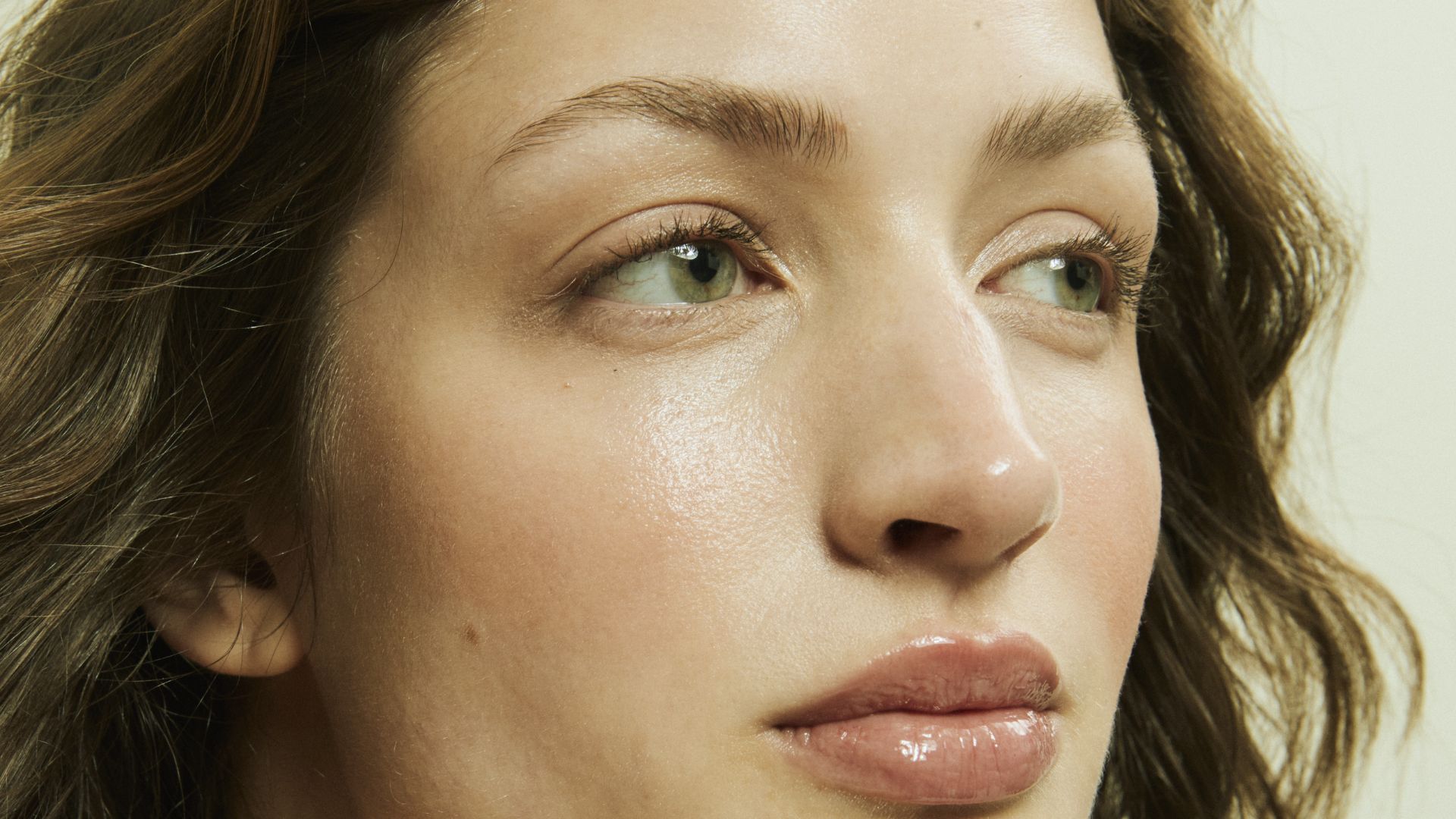 Close-up of a person with light green eyes gazing to the side, featuring smooth skin and wavy brown hair. The mood is serene and contemplative.