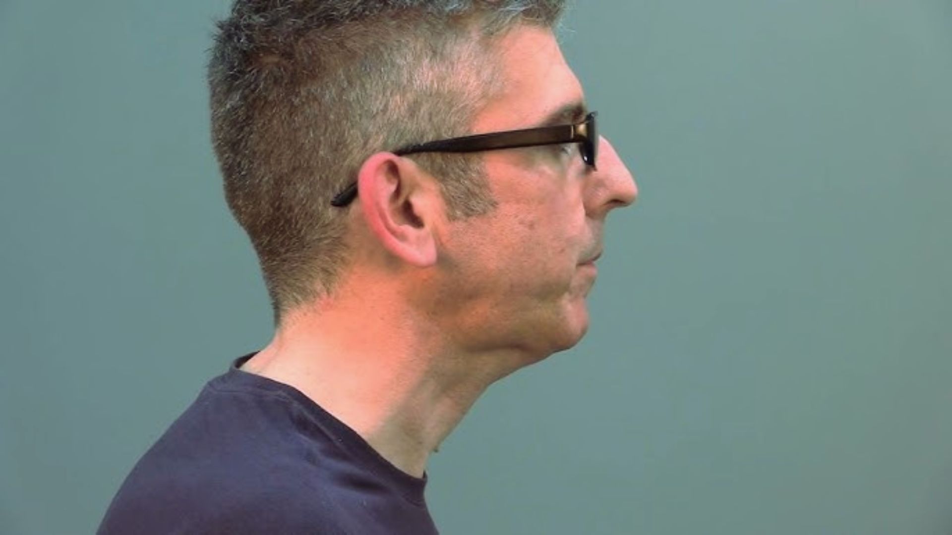 A man with short gray hair and glasses faces right against a plain teal background. His expression is neutral, and he wears a dark shirt.
