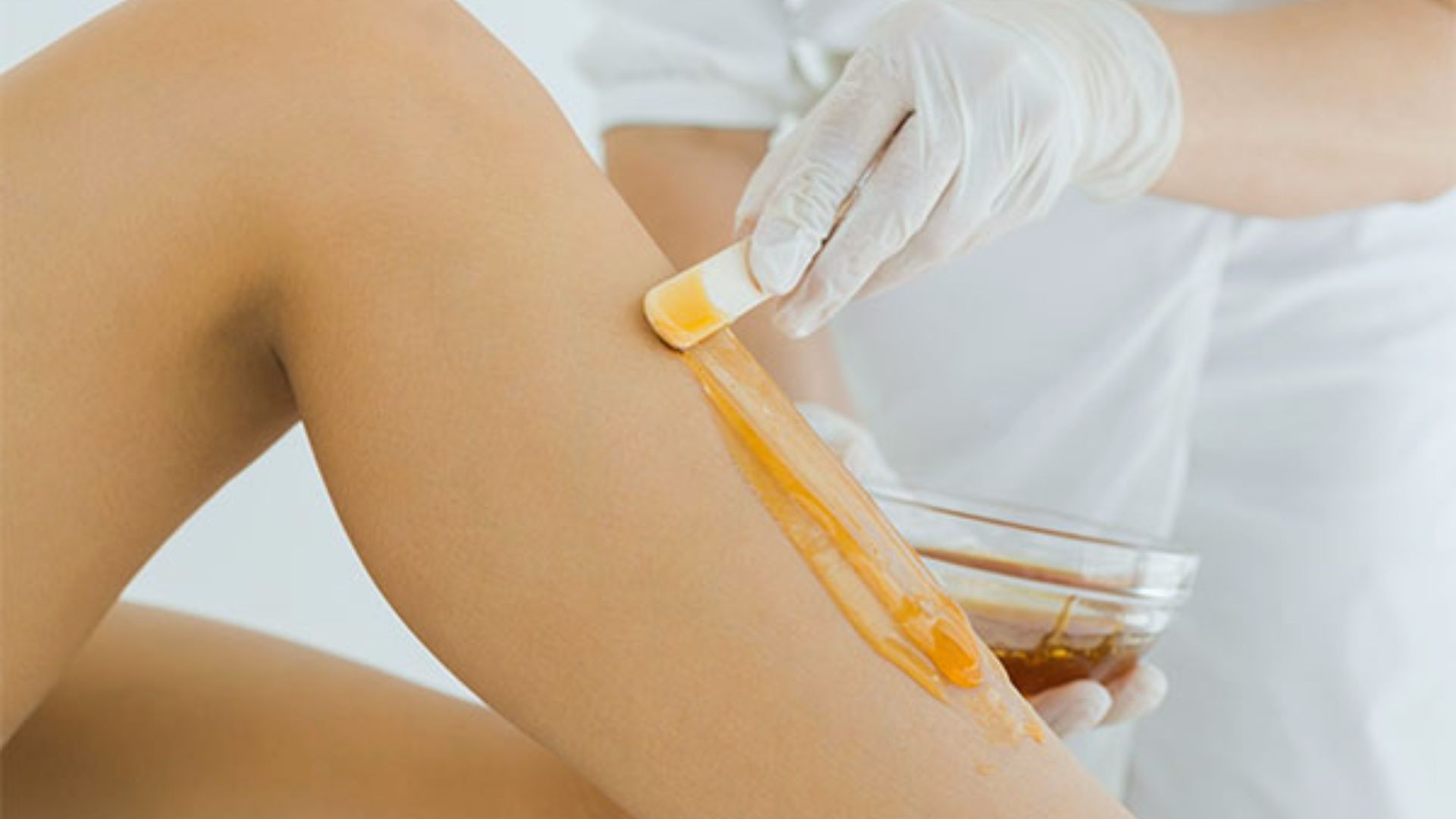 A person applies warm wax to another person's leg using a spatula in a spa setting. The professional wears gloves, emphasizing a clean, hygienic procedure.