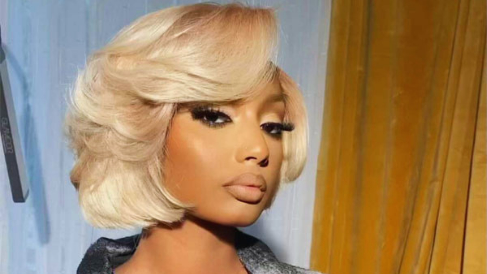 A person with a chic, short blonde bob and dramatic makeup stares confidently at the camera. The background features soft yellow and blue curtains.