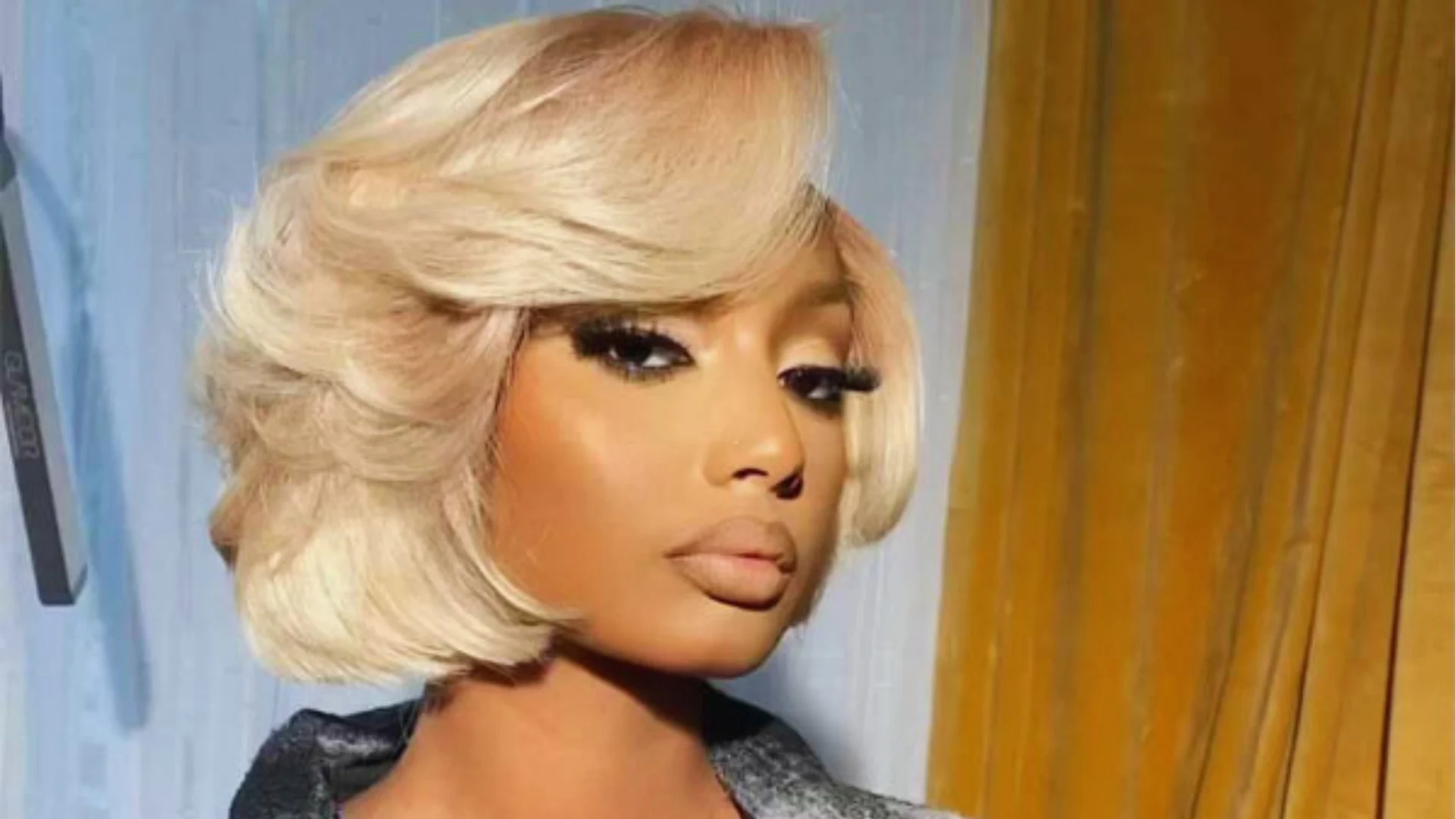 A person with a chic, short blonde bob and dramatic makeup stares confidently at the camera. The background features soft yellow and blue curtains.