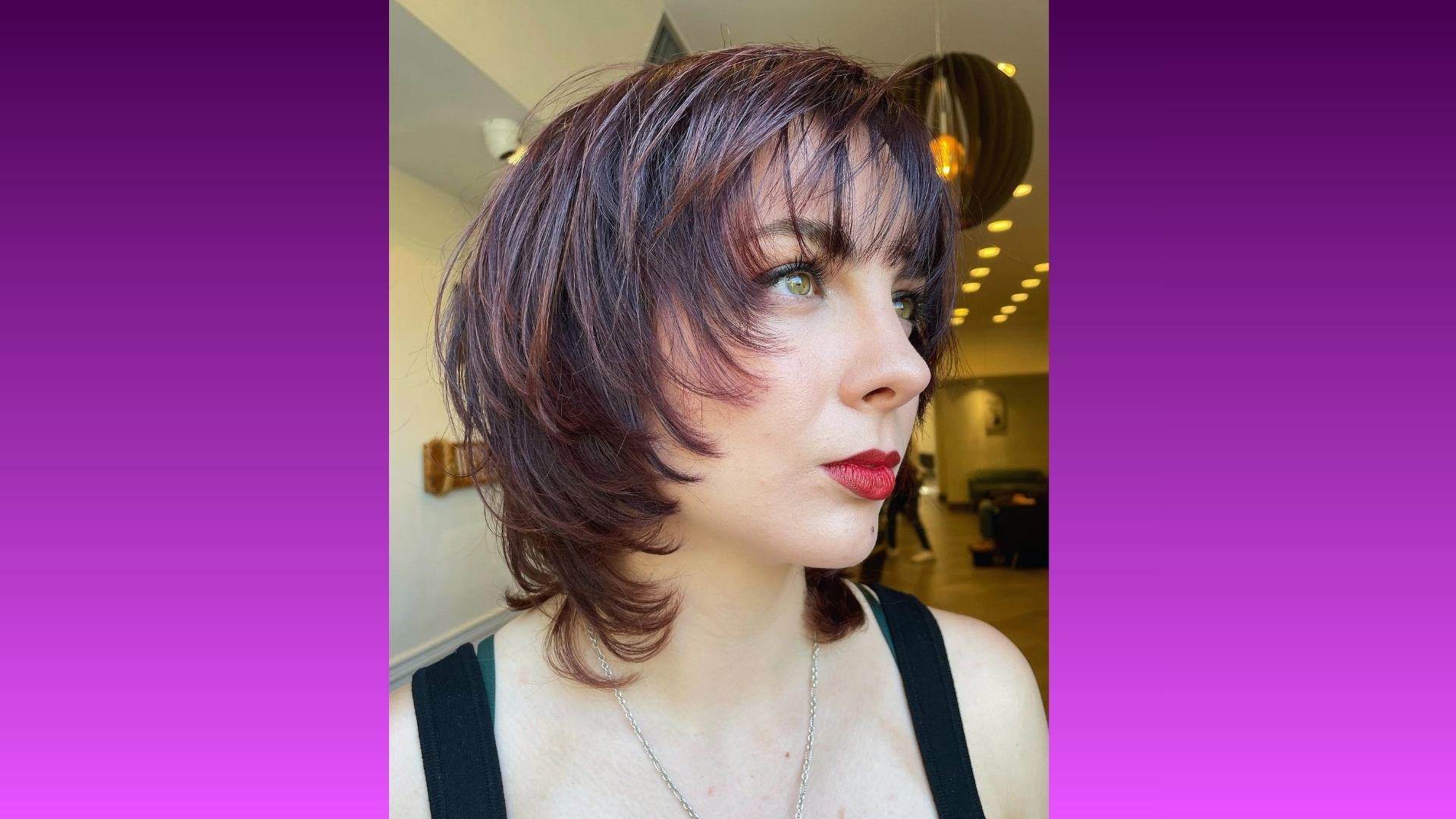 Woman with layered bob haircut and side-swept bangs looks to the side. She wears a black top and has red lipstick. Background is a modern room.