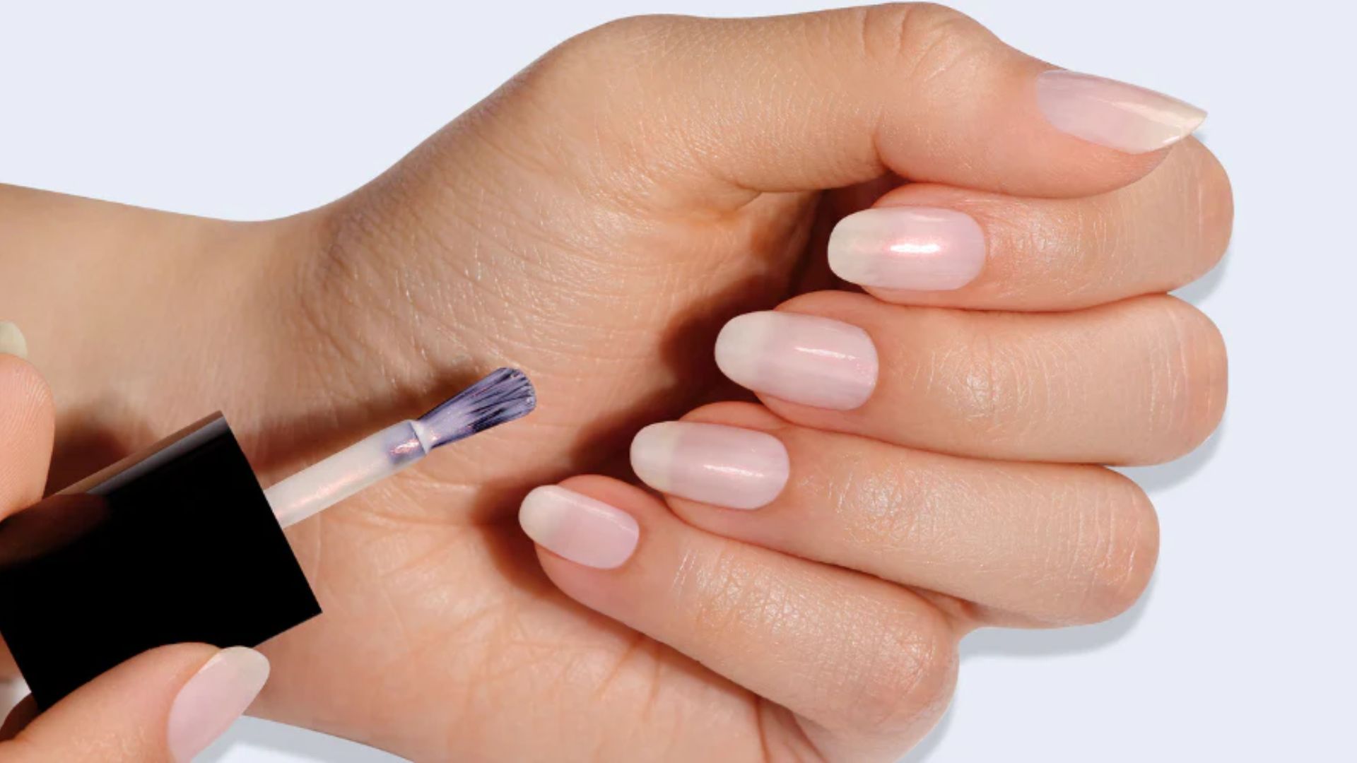 Close-up of a hand applying sheer pink nail polish to neatly manicured nails. The scene conveys a sense of care and personal grooming.