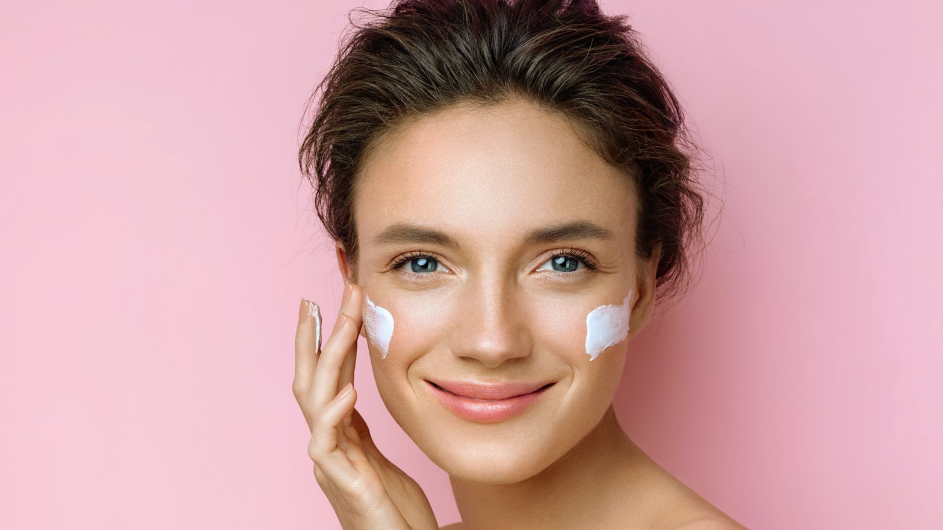 A smiling person with short brown hair applies cream to their cheeks against a pink background, conveying a sense of freshness and skincare.