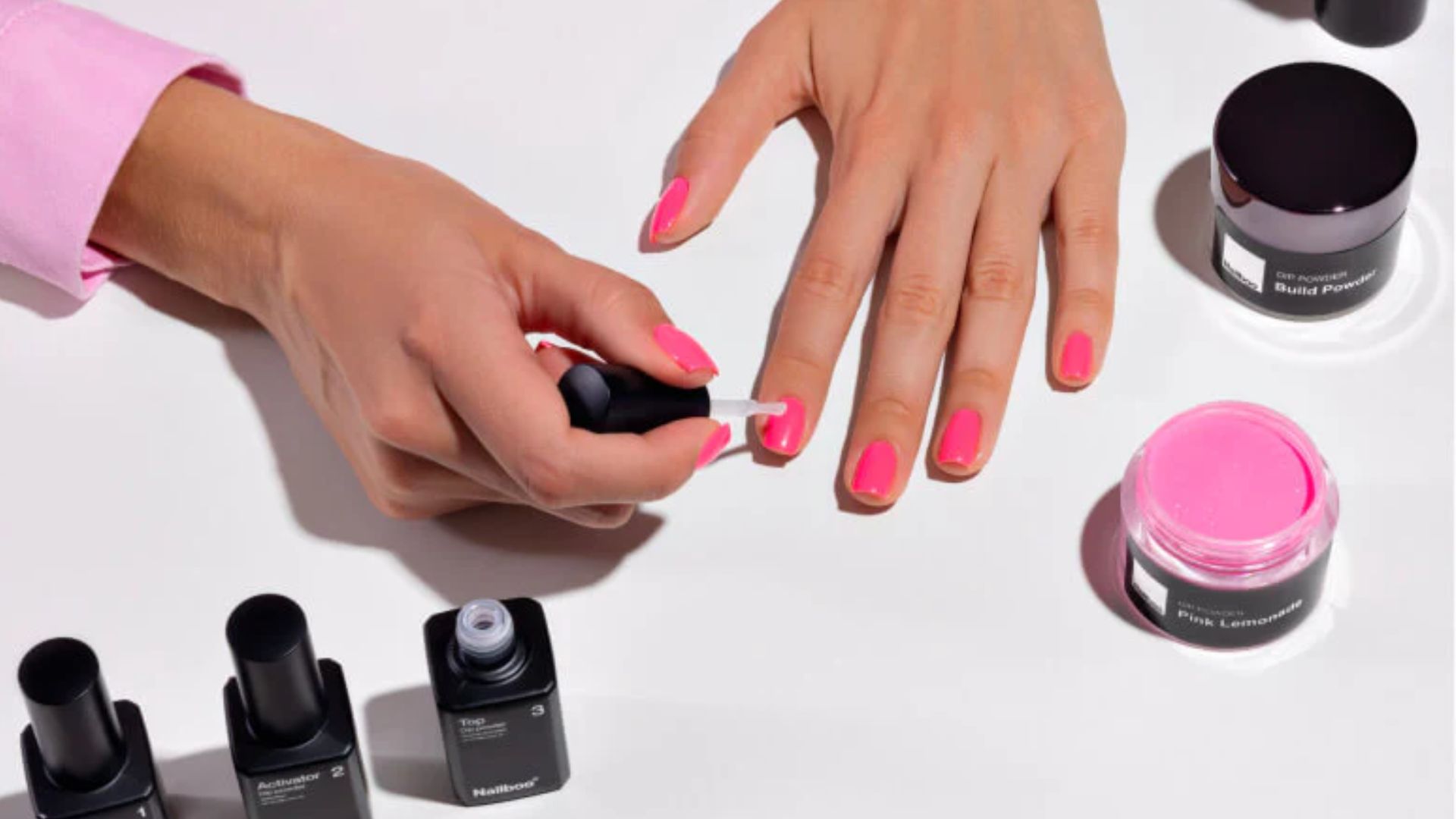 A hand applying bright pink nail polish to another hand on a white surface. Bottles of polish and a pink powder jar are scattered nearby, conveying a stylish, vibrant tone.