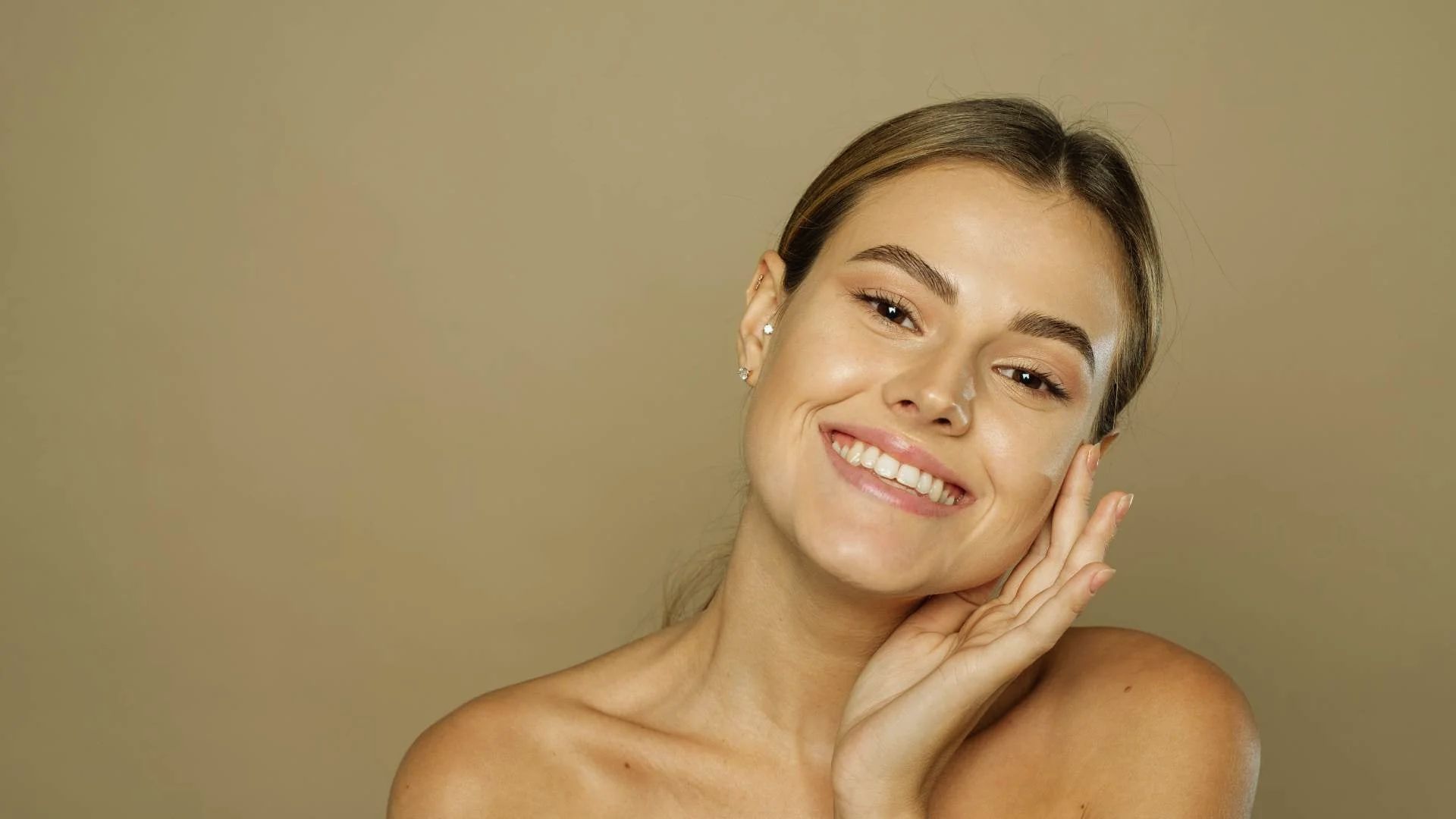 Smiling woman with smooth skin and natural makeup, resting her head on her hand against a beige background. Her expression is relaxed and content.