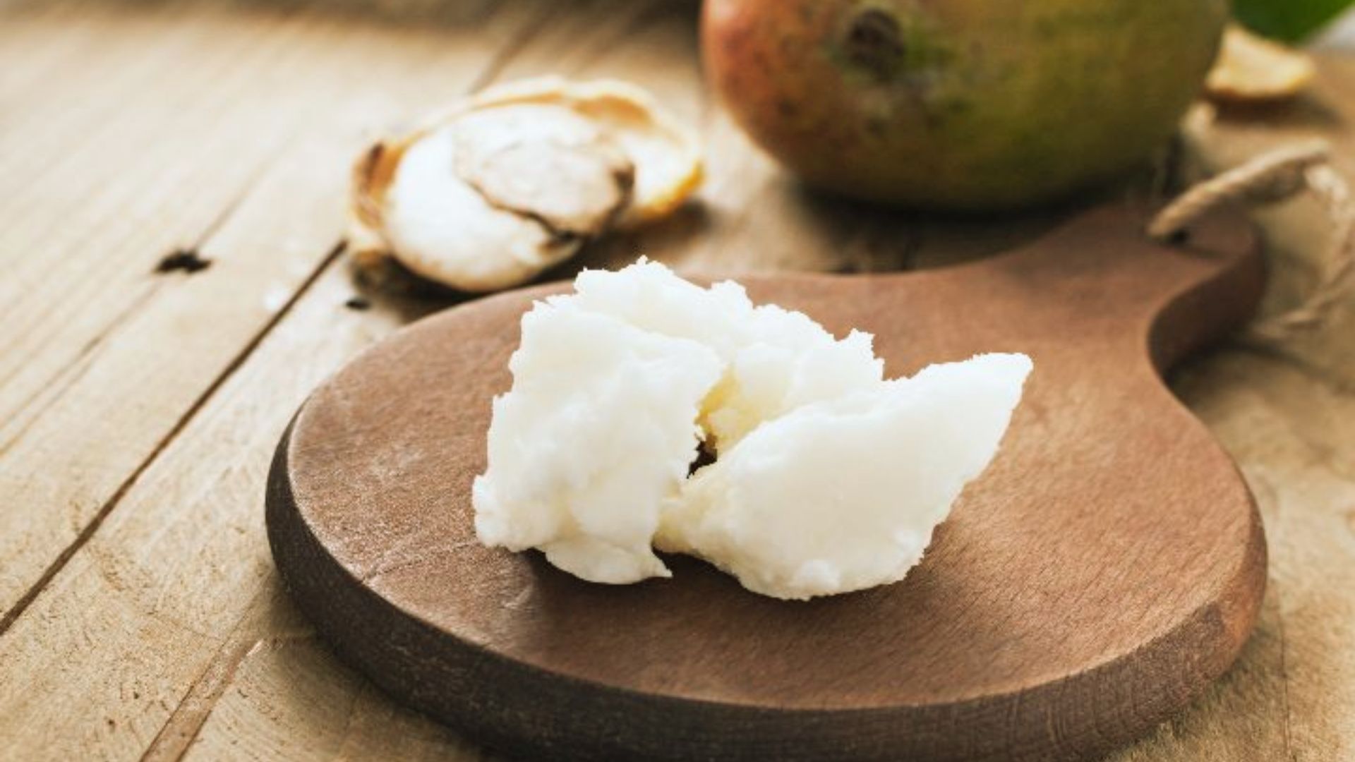 A wooden board with a dollop of white shea butter, set against a rustic wooden table. In the background, a mango and a cracked nut shell are visible.