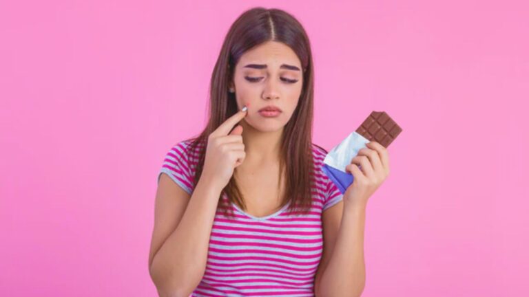 Woman with long hair frowns, holding chocolate in one hand and touching her cheek with the other. She wears a striped shirt against a pink background.