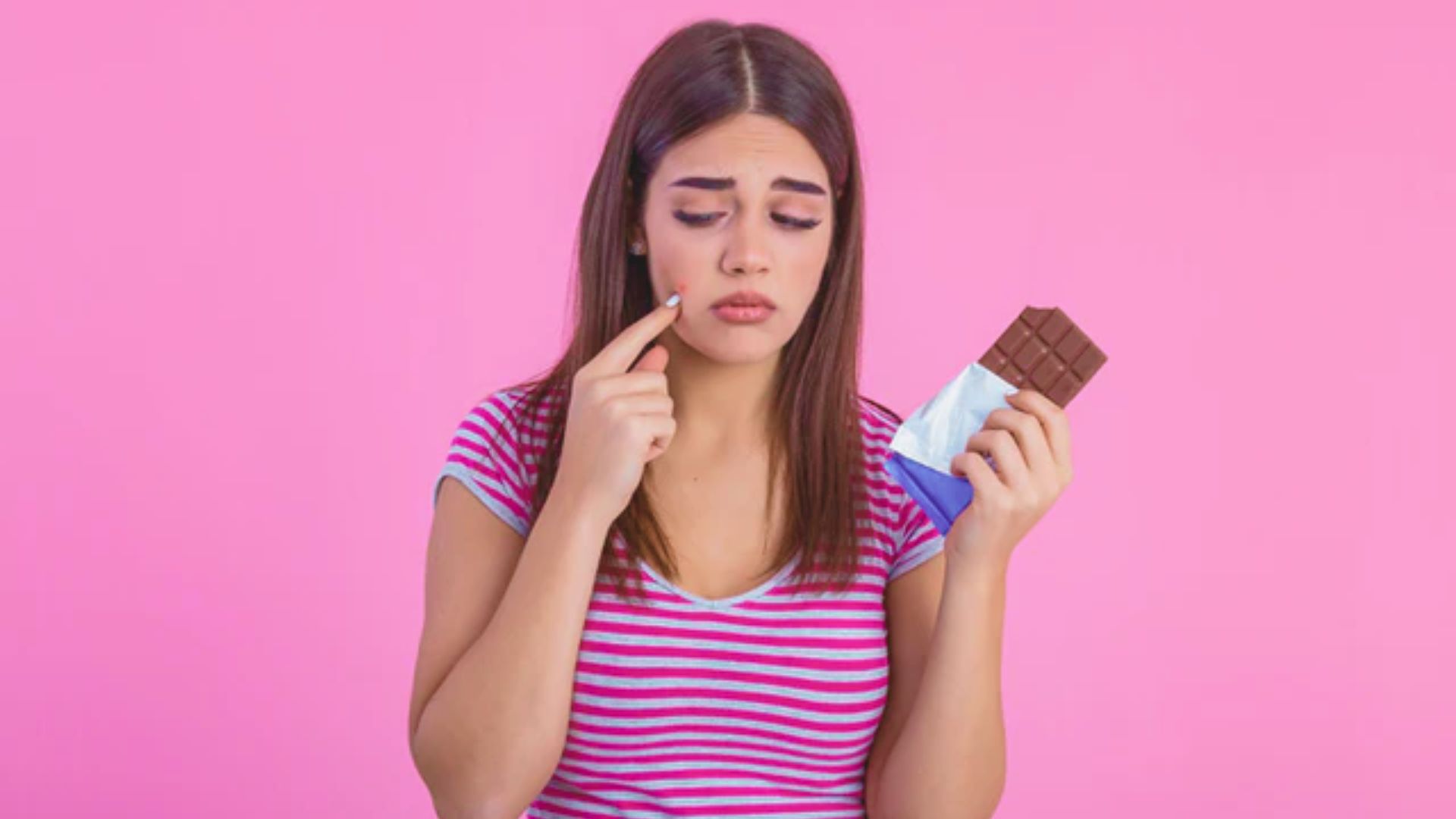 Woman with long hair frowns, holding chocolate in one hand and touching her cheek with the other. She wears a striped shirt against a pink background.