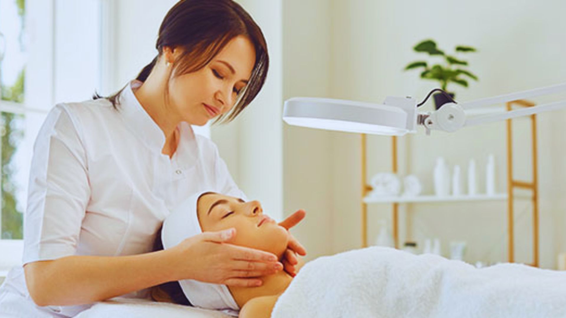 A woman gives a relaxing facial massage to a client lying under a white towel in a bright spa room. The atmosphere is calm and soothing.