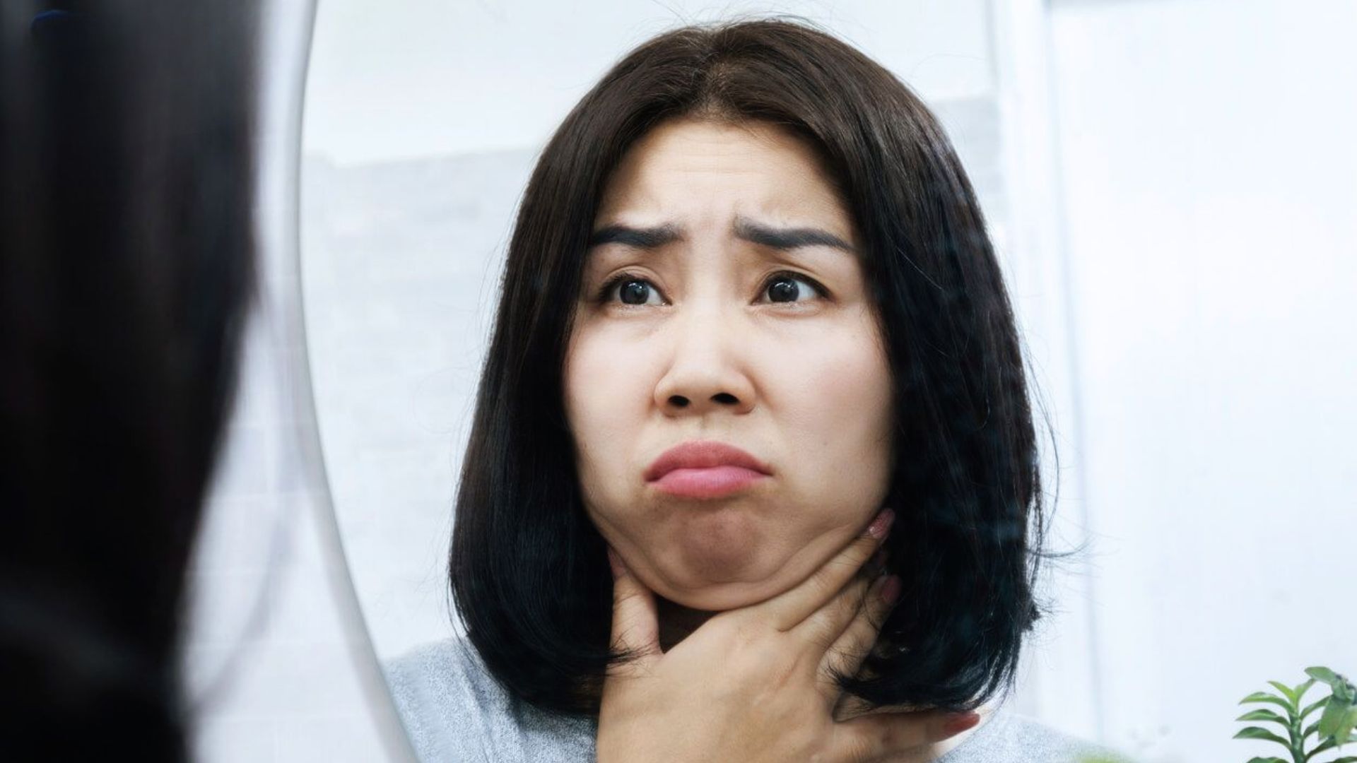 A woman with short dark hair looks into a mirror, holding her neck with both hands, appearing concerned. The room is brightly lit, suggesting a bathroom.