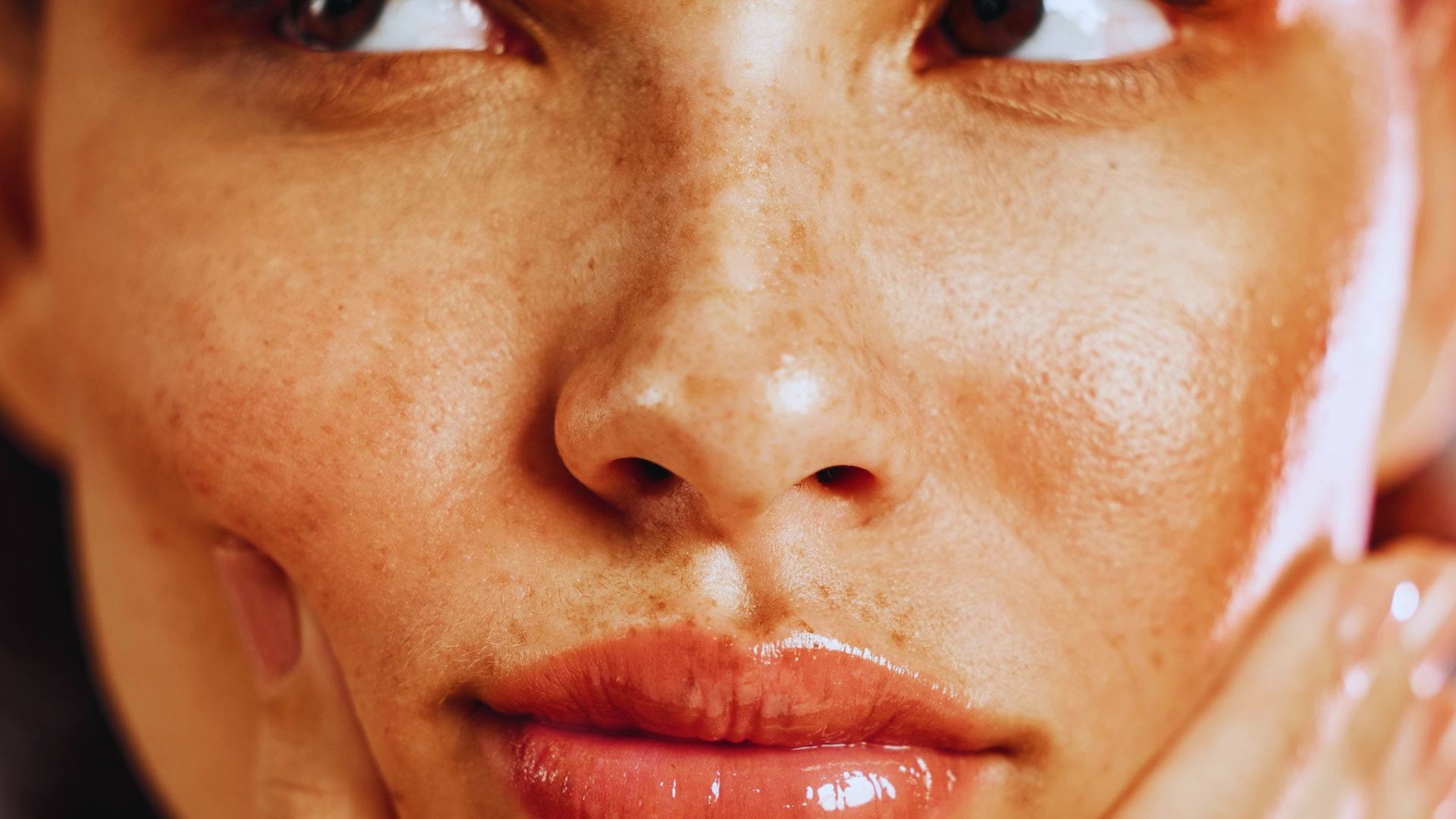 Close-up of a person's face with glossy skin and freckles. The person gently touches their cheek, conveying a sense of contemplation or relaxation.