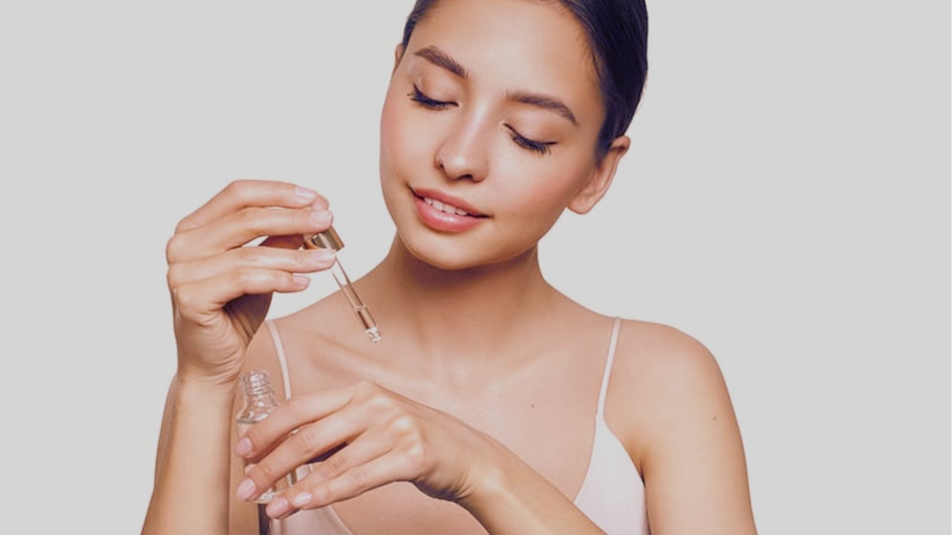 A woman with closed eyes gently applies serum to her skin using a dropper. She appears relaxed and content, conveying a sense of self-care.