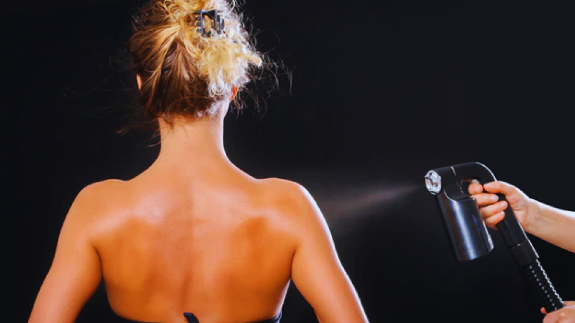 A person with tied-back hair receives a spray tan on their back, creating a smooth, even tone. The setting is against a black background, highlighting the process.