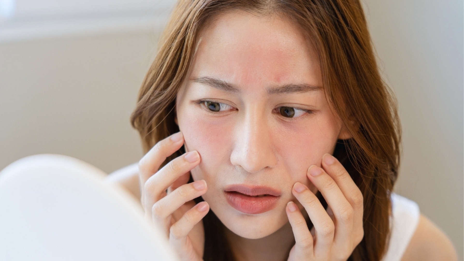 A woman looks concernedly at her reflection, touching her cheeks, which appear flushed and red. The setting is a softly lit room, conveying worry.