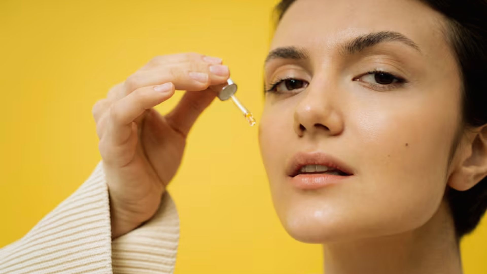 A person applies facial serum with a dropper against a vibrant yellow background. The scene conveys a sense of self-care and freshness.