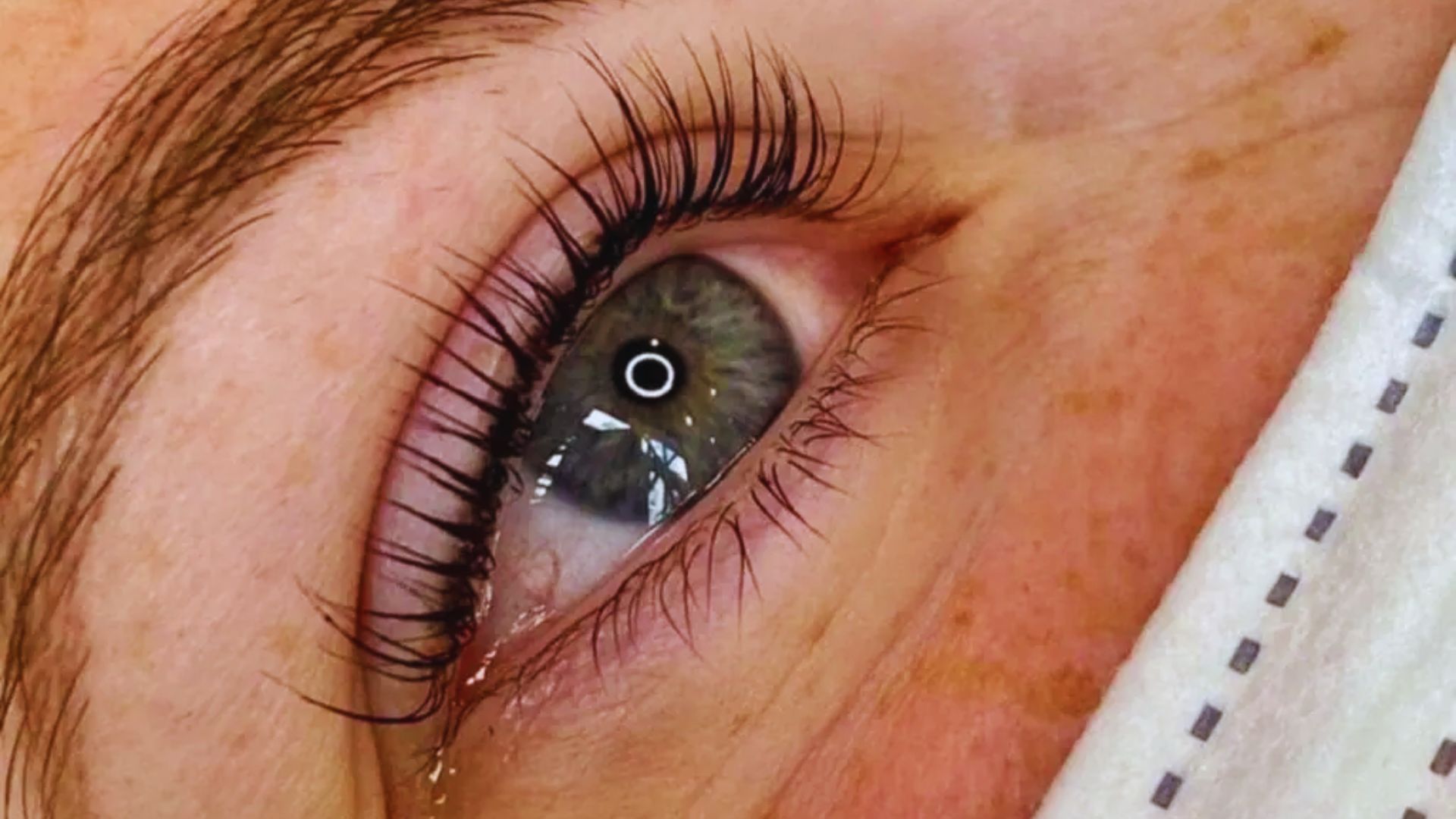 Close-up of a green eye with long, curved eyelashes. The skin is fair and freckled, and part of a white, patterned cloth is visible. The eye appears focused.