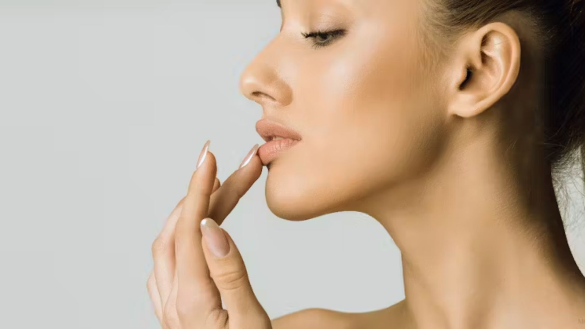 A woman with smooth skin gently touches her lips with her fingertips. Her serene profile is set against a soft gray background, conveying elegance.