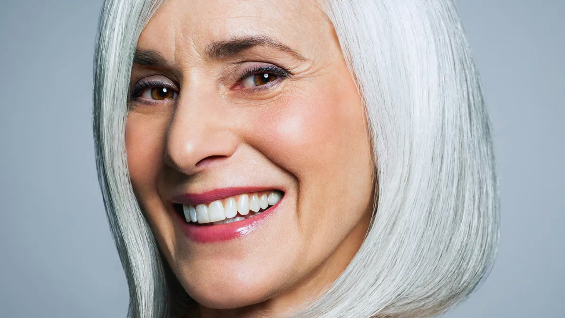 Smiling woman with shoulder-length white hair against a gray background. Her expression is joyful, conveying warmth and confidence.
