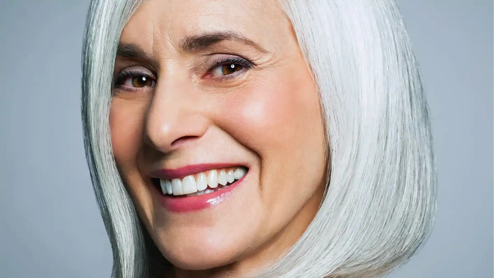 Smiling woman with shoulder-length white hair against a gray background. Her expression is joyful, conveying warmth and confidence.