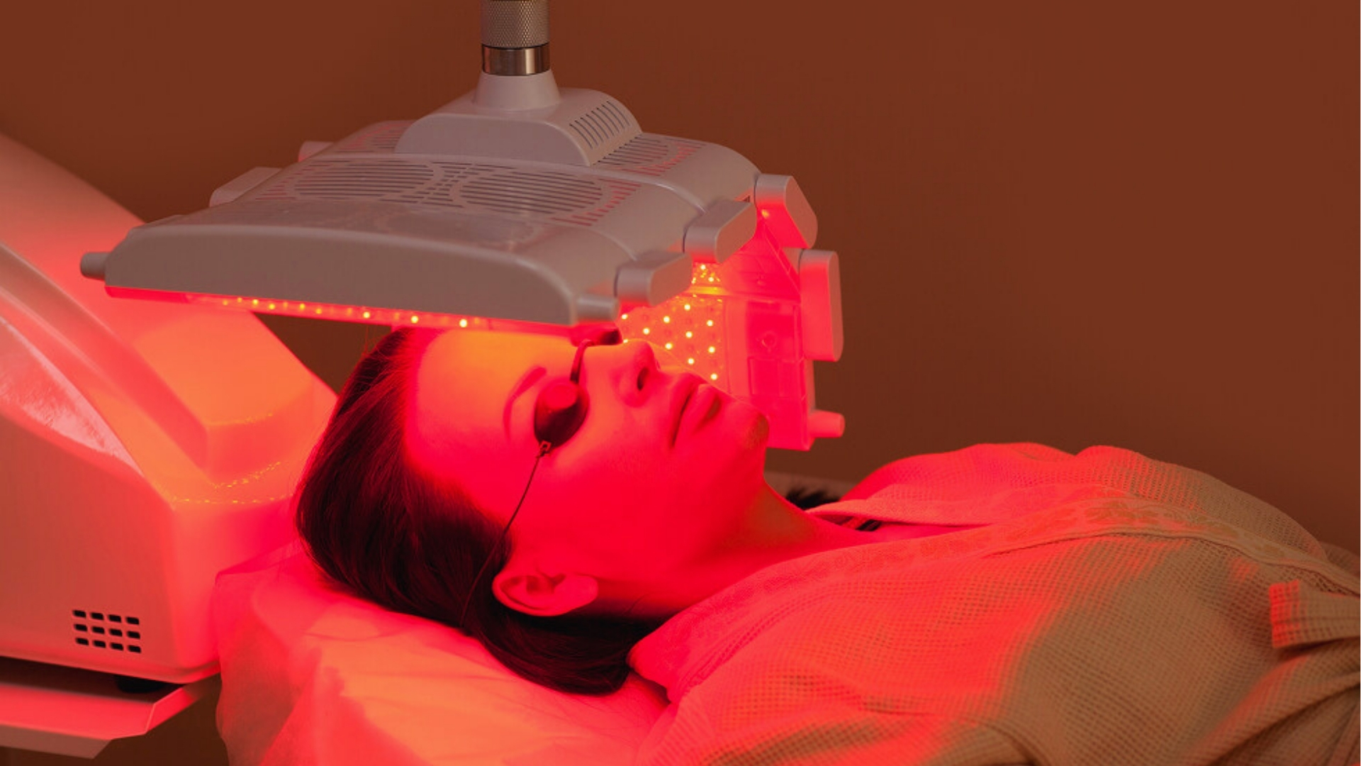 A woman lying under a red LED light therapy device, eyes covered with goggles. The serene setting suggests relaxation and self-care.